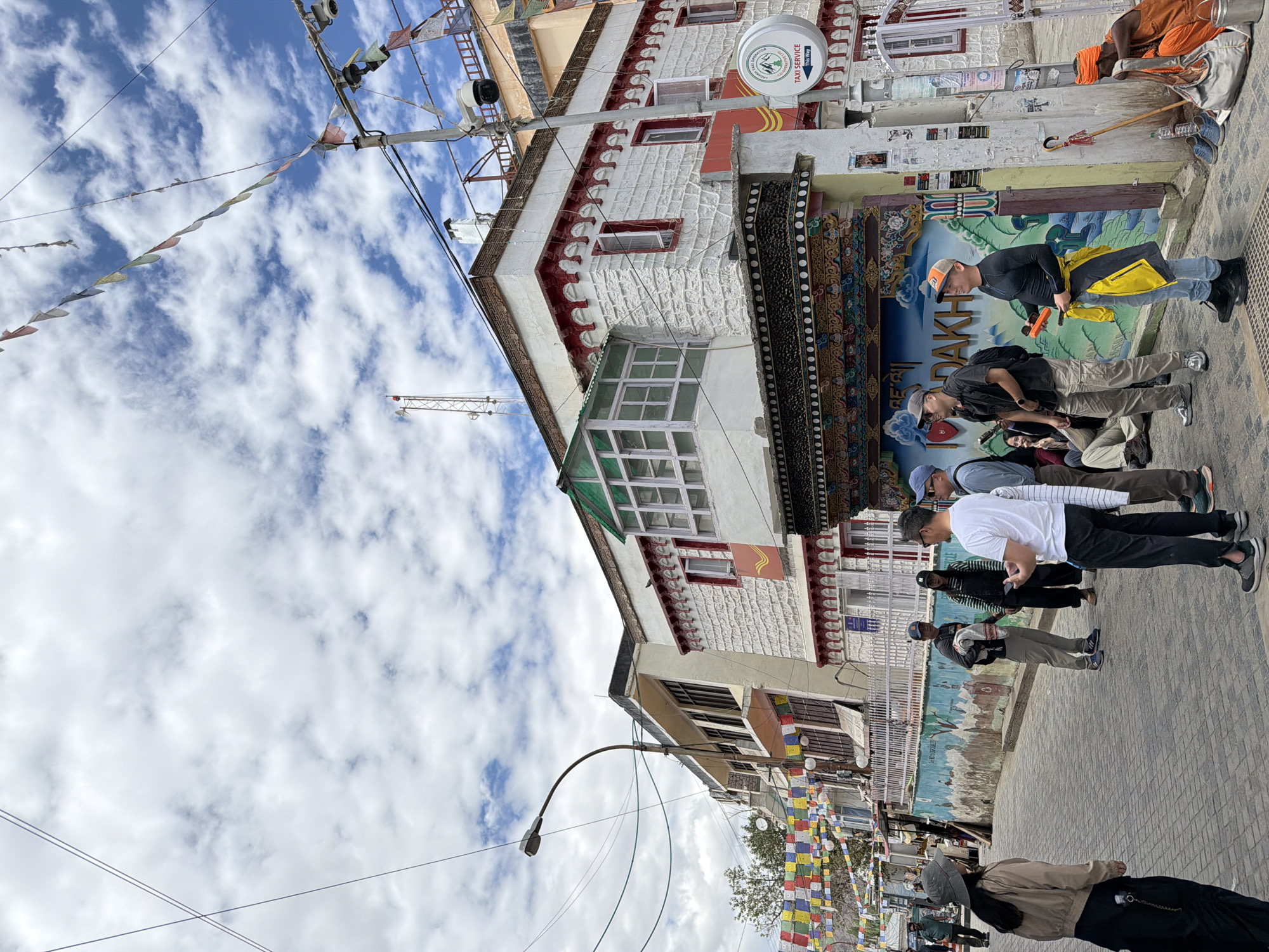 The image depicts a street scene in Leh, India, under a cloudy sky. In the foreground, a group of tourists are gathered, some looking at a mural on a building wall that reads "I Love Ladakh". The building itself is multi-story, with white textured walls and red decorative trim. Prayer flags are strung across the street. Further down the street, a sadhu (holy man) in orange robes sits on the ground next to a bag and a bucket. Other people are seen walking and standing in the street and on sidewalks. The street is paved with cobblestones. Utility poles with wires and cameras are visible.