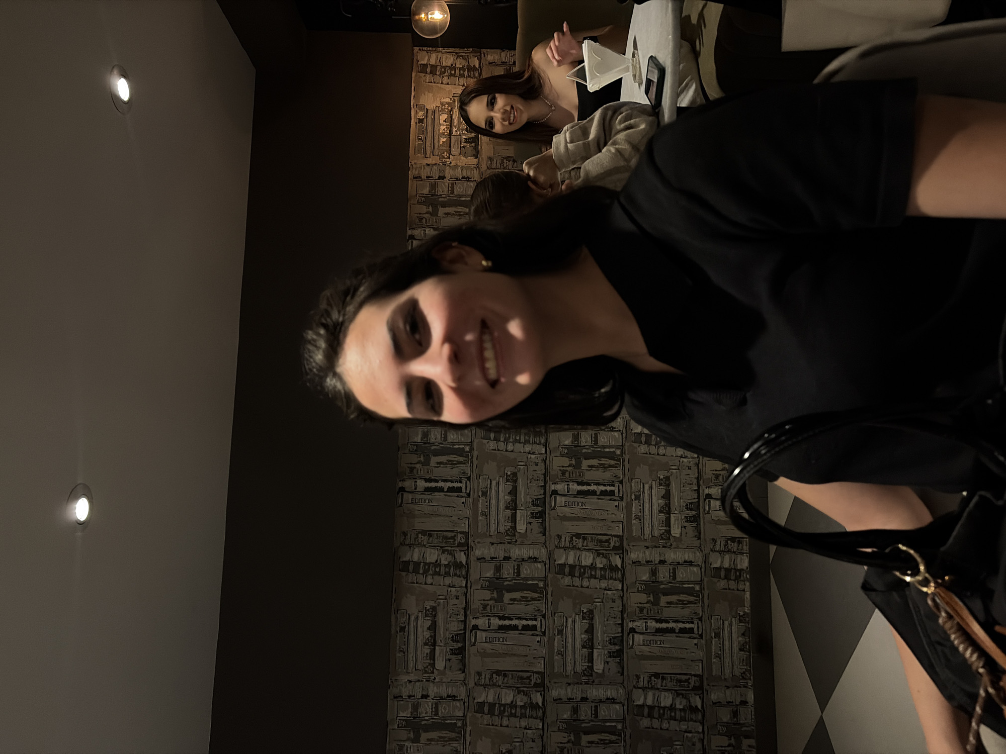 The scene is an indoor establishment, likely a cafe or lounge, located in Puebla, Mexico, as per provided information. In the foreground, a young woman with dark hair, wearing a black collared shirt and small gold hoop earrings, smiles directly at the camera; her face is slightly blurred. She holds a black handbag with a patent leather strap. To her right, a second individual, partially obscured from behind, shows the back of their dark hair and a light-colored sweater sleeve. In the background, a third young woman with long dark hair, wearing a black strapless top and a pearl necklace, is seated at a table, also smiling towards the camera. On her table rests a white, ridged, conical pour-over coffee device and a black smartphone. The wall behind the subjects is covered with wallpaper depicting grayscale stacks of books. The dark gray ceiling contains recessed spotlights. A spherical pendant light with an amber-tinted bulb hangs on the far right. The floor features a checkered pattern of dark gray and light-colored tiles.