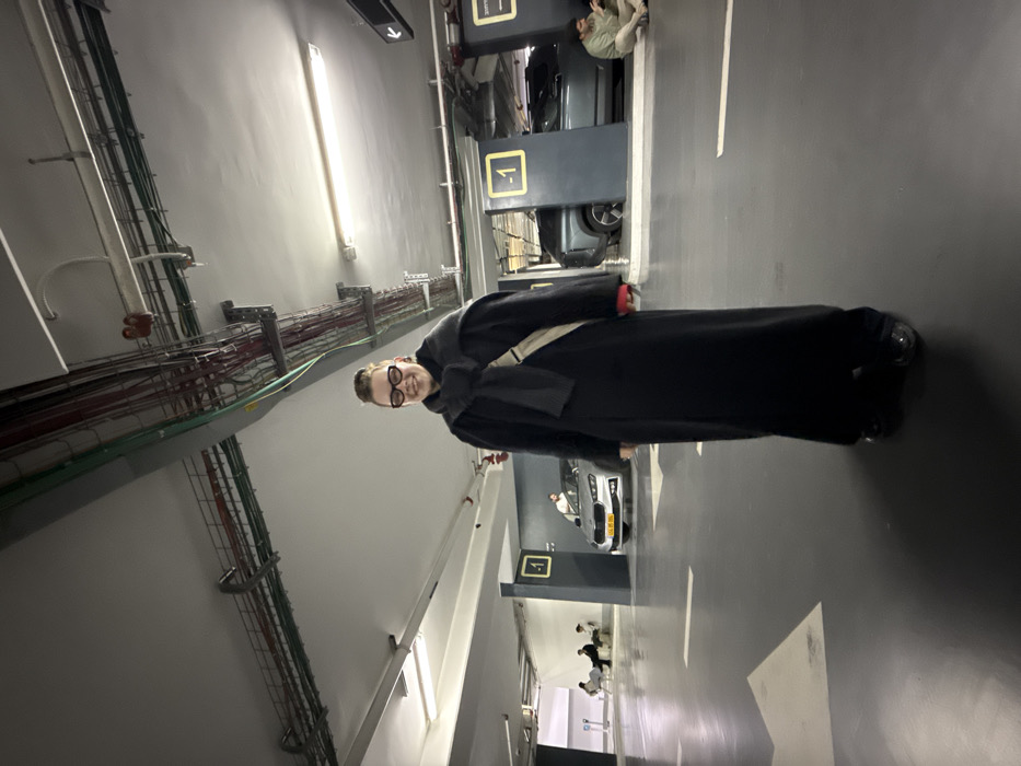 An individual stands centrally in an underground parking garage in Tel-Aviv, Israel, facing the camera with a slight smile. The person wears a long, dark (likely black) coat reaching the ankles, a dark scarf draped around the neck and shoulders, and a light-colored crossbody bag. Dark-rimmed glasses are worn, and red cuffs are visible at the wrists. The floor is dark gray with white directional arrows and lines.
In the background to the left, three individuals are seated on a white bench against a light gray wall. A silver Skoda Fabia with an Israeli yellow license plate is parked behind them, next to a dark gray pillar marked with a yellow "-1" sign. Further to the right, a dark gray SUV is parked, partially obscured by another dark gray pillar also marked with "-1." An individual is seated on the floor next to this SUV, looking down at a device.
The ceiling consists of exposed concrete, crisscrossed by numerous utility pipes, conduits, and cables in various colors (red, green, gray), along with fluorescent light fixtures and red fire suppression system components. The overall lighting is artificial and uniform.