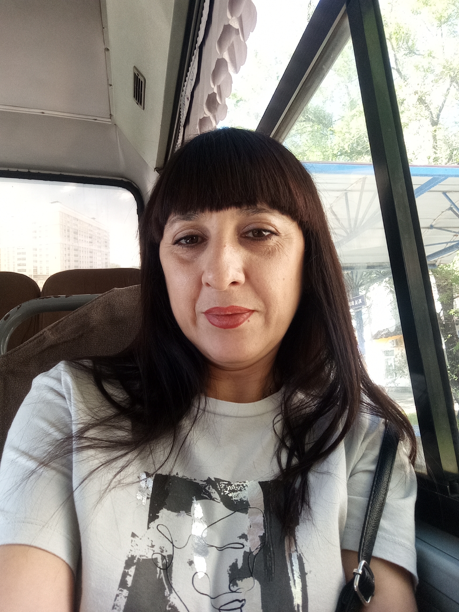 The image shows a medium shot of a woman with dark brown hair and bangs, wearing a white t-shirt with a black and white graphic print. She has dark eyes, noticeable wrinkles around her eyes, and is wearing red lipstick. She is sitting inside a vehicle, likely a bus or train, with a window on her right side. Through the window, trees and a building can be seen in the background. The interior of the vehicle is visible, including a grey upholstered seat behind her and part of the ceiling. A black handbag with a silver buckle rests on her right shoulder. The scene appears to be captured during daylight. The specific location in the city of Belogorsk, Russia is not discernible from the visual information provided in the image.