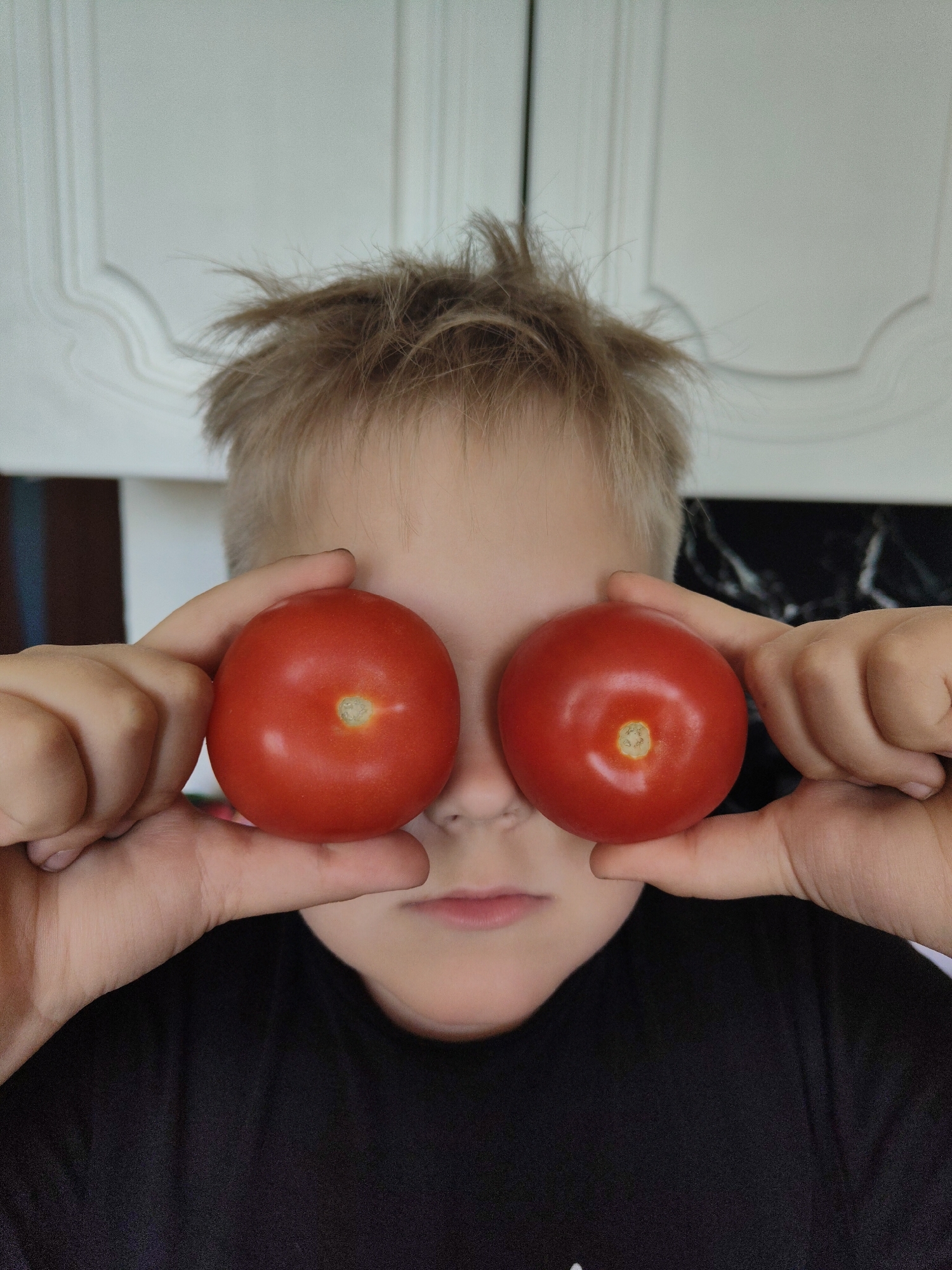 A fair-skinned child, with short, light brown hair, is holding two red tomatoes up to their eyes, obscuring them from view. The child is wearing a black shirt. The background consists of white cabinets with detailed moldings and dark, possibly granite countertops. The location is inside, likely a kitchen. The city and exact location are unknown.