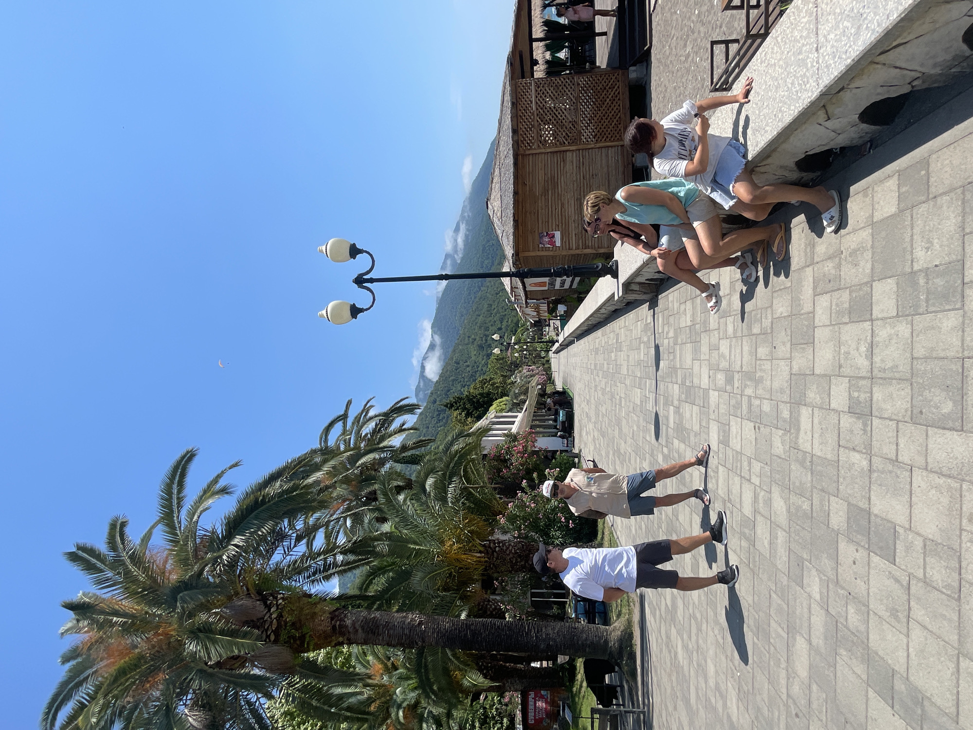 The image depicts a sunny day along a paved promenade in Abaata, Abkhazia. Tall palm trees line the left side of the walkway, with buildings and open-air gazebos visible further down. In the background, lush green mountains rise towards a clear blue sky, with a few clouds obscuring the peaks.
On the promenade, two men in casual attire are standing and talking. One man wears a white t-shirt, dark shorts, and a black baseball cap, while the other is in a sleeveless beige shirt, light shorts, and a white cap. Further along, a group of four people are seated on a low stone wall. Three women are visible: one in a light blue tank top and shorts, another in a black tank top and shorts, and a younger person in a white t-shirt and denim shorts. A fourth person, partially obscured, is also seated. To the right of the promenade, a wooden gazebo with a thatched roof stands on the sand, suggesting it is near the beach. A single street lamp with two lights is prominent on the left side of the walkway. A paraglider is visible in the sky above the mountains.