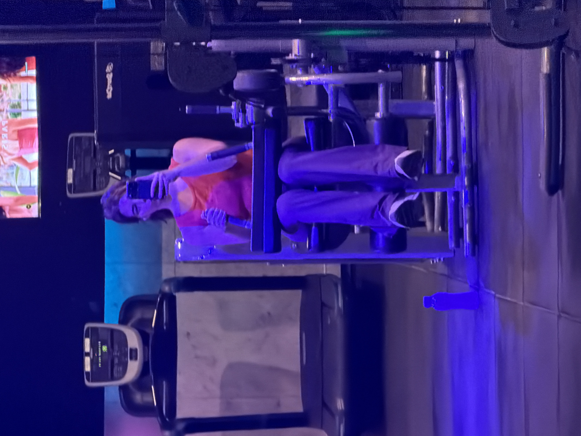A person is seated on a gym exercise machine, likely a leg press, in a dimly lit fitness center. The individual, appearing to be a woman, is wearing an orange tank top and dark trousers. She is holding a smartphone in her right hand, seemingly taking a selfie or recording herself, with her left hand resting on the machine's handle. Her hair is dark and falls around her face.
The gym features dark blue and purple ambient lighting, creating a cool and moody atmosphere. In the background to the far left, a large screen displays an image or video, where the word "SYBARIS" is partially visible. Another gym equipment console, possibly from a treadmill or elliptical, is in the foreground at the bottom left, showing some unreadable text and icons on its display. A blue water bottle lies on the floor to the right of the seated person. The wall behind her appears to be tiled or textured. The machine she is using has a partially visible "Technogym" logo. The scene depicts an indoor fitness activity, possibly during evening hours given the artificial lighting. No specific visual cues indicate the location is Buenos Aires, Argentina. The person's expression is neutral and focused on her phone.