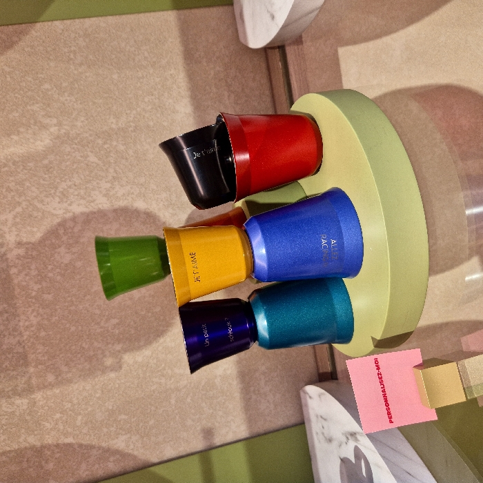 The image displays a vibrant collection of small, colorful cups or containers arranged on a light green, circular stand. The perspective is an overhead, slightly angled view, with the overall image rotated clockwise.
In the center of the frame, seven individual cups are neatly placed around the edge of a flat, light green circular base. These cups are made of a metallic or glossy material, reflecting some light. They are arranged in a semi-circular or U-shaped pattern.
From left to right (as the cups are arranged in the image):
1. A bright green cup, slightly wider at the top.
2. A shiny golden-yellow cup. Text visible on its side reads "JE T'AIME" (I love you).
3. A deep purple cup. Text visible on its side reads "Un petit chouchou?" (A little darling/favorite?).
4. A teal or dark turquoise cup.
5. A sparkling blue cup. Text partially visible on its side reads "ALLEZ FAÇ...".
6. A bright red cup.
7. A black cup, which appears to be nested inside the red cup, with its rim slightly protruding. Text visible on its side reads "Je t'aime".
The light green circular stand provides a foundation for the cups. This stand is situated on a textured, light brown surface, which could be a floor or a large table. The surface has subtle variations in shade, suggesting a rough or patterned texture.
In the bottom right corner of the image, there is a small, rectangular pink note. Red text printed on it clearly reads "PERSONNALISEZ-MOI" (Personalize me). Adjacent to this note, to its left, is a small, golden, cube-like object.
In the background, to the top and bottom of the frame, parts of other structures are visible. To the top-left, there's a smooth, pale green surface. To the bottom-right, there are elements that appear to be made of white marble with gray veining, possibly decorative objects or parts of a display. Shadows are cast by the cups and the circular stand onto the brown surface, indicating an ambient indoor light source.
The scene suggests a display of customizable items, possibly in a retail store or an exhibition, emphasizing personalization and affection.