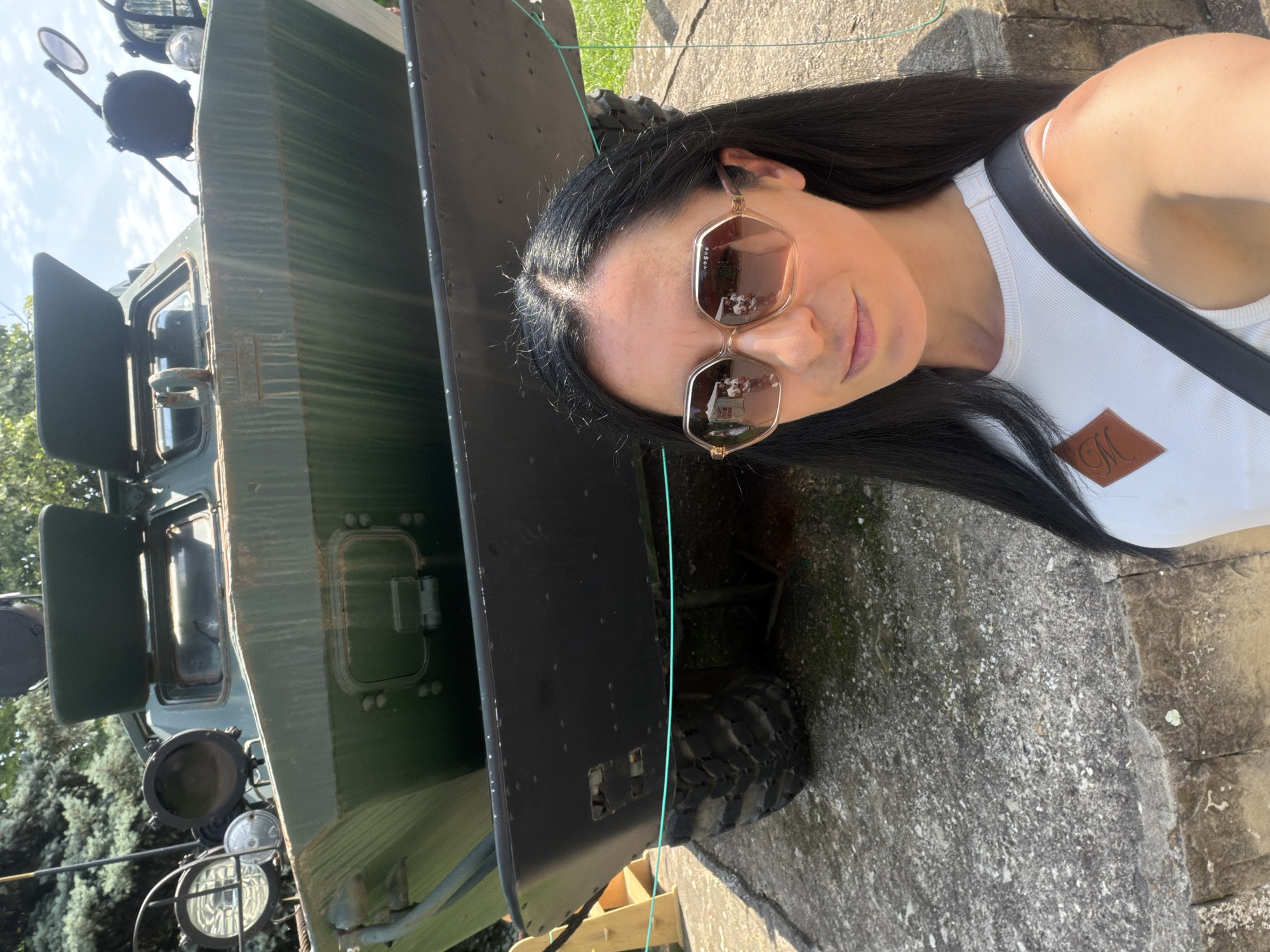 A woman takes a selfie in front of a weathered green military armored personnel carrier, likely a BTR-series vehicle, outdoors in Pleven, Bulgaria. The woman, positioned in the foreground, has long dark hair and wears oversized hexagonal sunglasses with "VOGUE" branding. Reflections in her sunglasses show two figures and white structures. She wears a white ribbed tank top with a brown diamond-shaped patch featuring an embossed 'M' and a black strap over her right shoulder.
The military vehicle dominates the mid-ground, displaying a dull olive-green color with visible streaks and signs of wear. Its front features large circular headlights and a smaller auxiliary light on the left side, a partially open top hatch, and a dark protective bumper across the lower front. A robust tire with a deep tread pattern is visible on the right side of the vehicle, resting on a concrete surface. A thin, light green rope acts as a barrier in front of the vehicle.
The ground consists of weathered concrete or stone pavement. In the background, dense green foliage from trees is visible, along with a clear blue sky and scattered white clouds, indicating bright, direct sunlight. The scene suggests a military museum or memorial park setting.