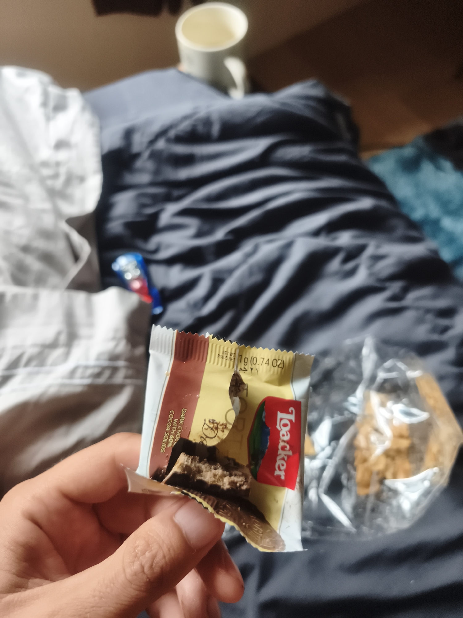 A close-up, eye-level indoor shot shows a person's hand holding a partially opened package of a Loacker dark chocolate snack. The snack bar inside the wrapper is partly eaten, revealing layers of dark chocolate and wafer. The packaging displays text including "Loacker", "DARK CHOC WITH 60% COCOA SOLIDS", and "1g (0.74 OZ) POIDS NET". To the right of the hand, another clear plastic bag contains more similar snacks. In the blurred background, there are rumpled bedsheets in light grey/white and dark blue/grey tones. A white mug is visible in the upper background, and a small blue and red object (possibly a lighter or a small toy) is on the bedding further back. The overall scene suggests a casual moment, possibly relaxing in bed while having a snack, in Baan Mae Nam, Thailand.