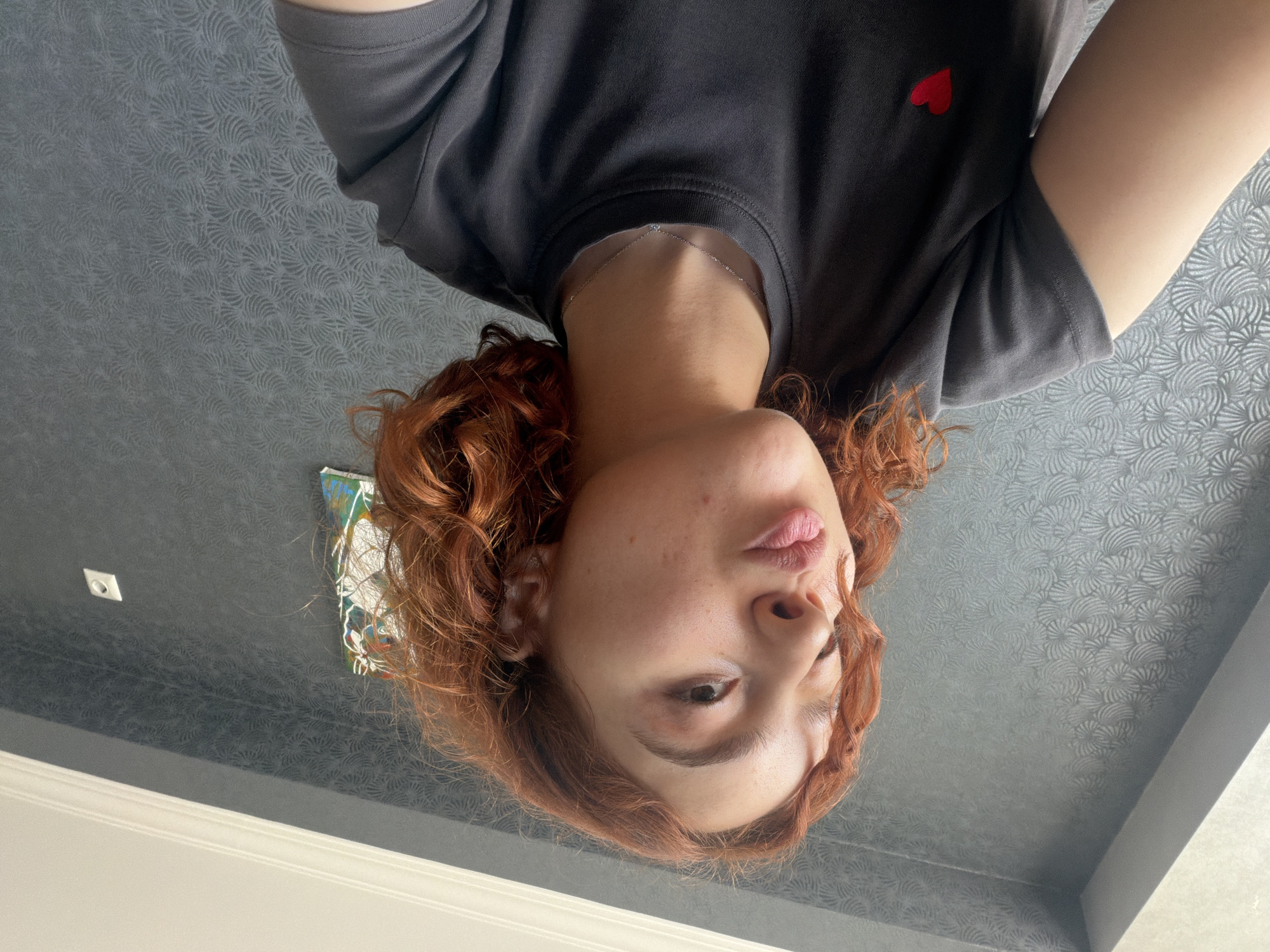 A person with red curly hair is facing the camera in a selfie taken at a low angle. The person is wearing a gray t-shirt with a small red heart on the chest. The background is a gray wall with a subtle patterned design. In the top left corner, there's a light switch and part of a painting. The location is likely indoors, but there is no clear indication of it being in Tbilisi, Georgia.