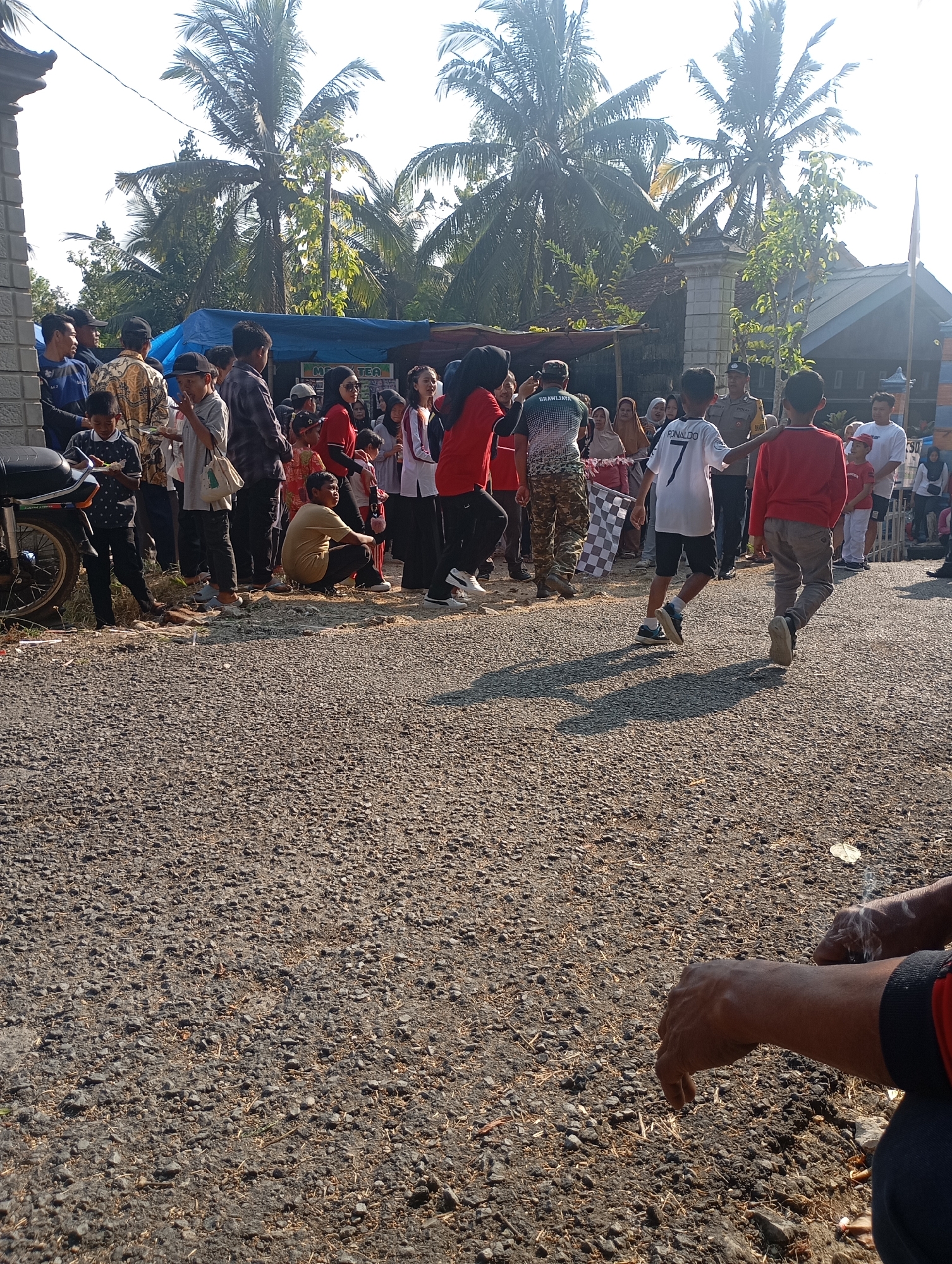 A group of people are gathered outdoors in what appears to be a village or community event in Ketepung, Indonesia. The scene is likely a street or open area, with a gravel ground surface. In the background, there are palm trees and traditional Indonesian buildings with dark roofs. Several individuals, including children and adults, are present, some standing, some sitting, and some walking.
There are a few notable elements within the crowd. Two young boys are walking away from the camera, one wearing a white shirt with "RONALDO" and the number 7 on the back, and the other in a red shirt. A checkered flag is being held by someone in the background. A man in a police uniform is visible interacting with the crowd. There are also tents or stalls set up, suggesting a vendor or event setup. A motorcycle is parked on the left side of the image. The overall atmosphere seems to be one of a casual outdoor gathering or local celebration. The sunlight indicates it is daytime.