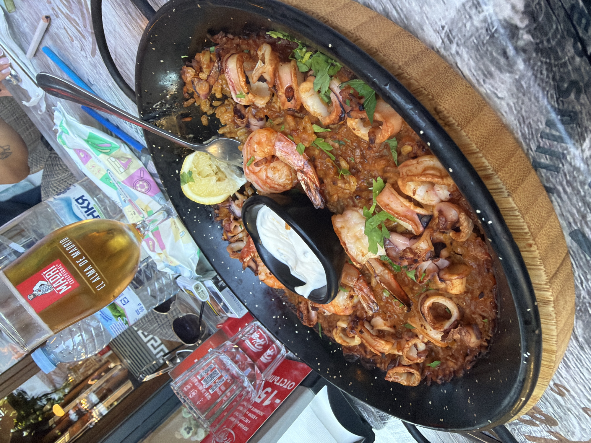 A close-up, high-angle shot features a large, black paella pan filled with seafood and rice, positioned on a wooden table. The paella contains prawns, squid rings, and mussels, garnished with fresh parsley and a lemon wedge. A small black bowl of white sauce, likely aioli, sits in the center of the dish. A fork is submerged in the paella, suggesting it is being eaten.
In the background, a partially visible glass of beer with a "Madri" label is tilted. Bottles of water and other items are scattered on the table, including a red Coca-Cola glass and a pack of cigarettes. The table surface appears to be made of distressed wood. The overall ambiance suggests a casual dining experience, likely at a restaurant in Burgas, Bulgaria, given the presence of the paella, a Spanish dish, and the style of the setting.
