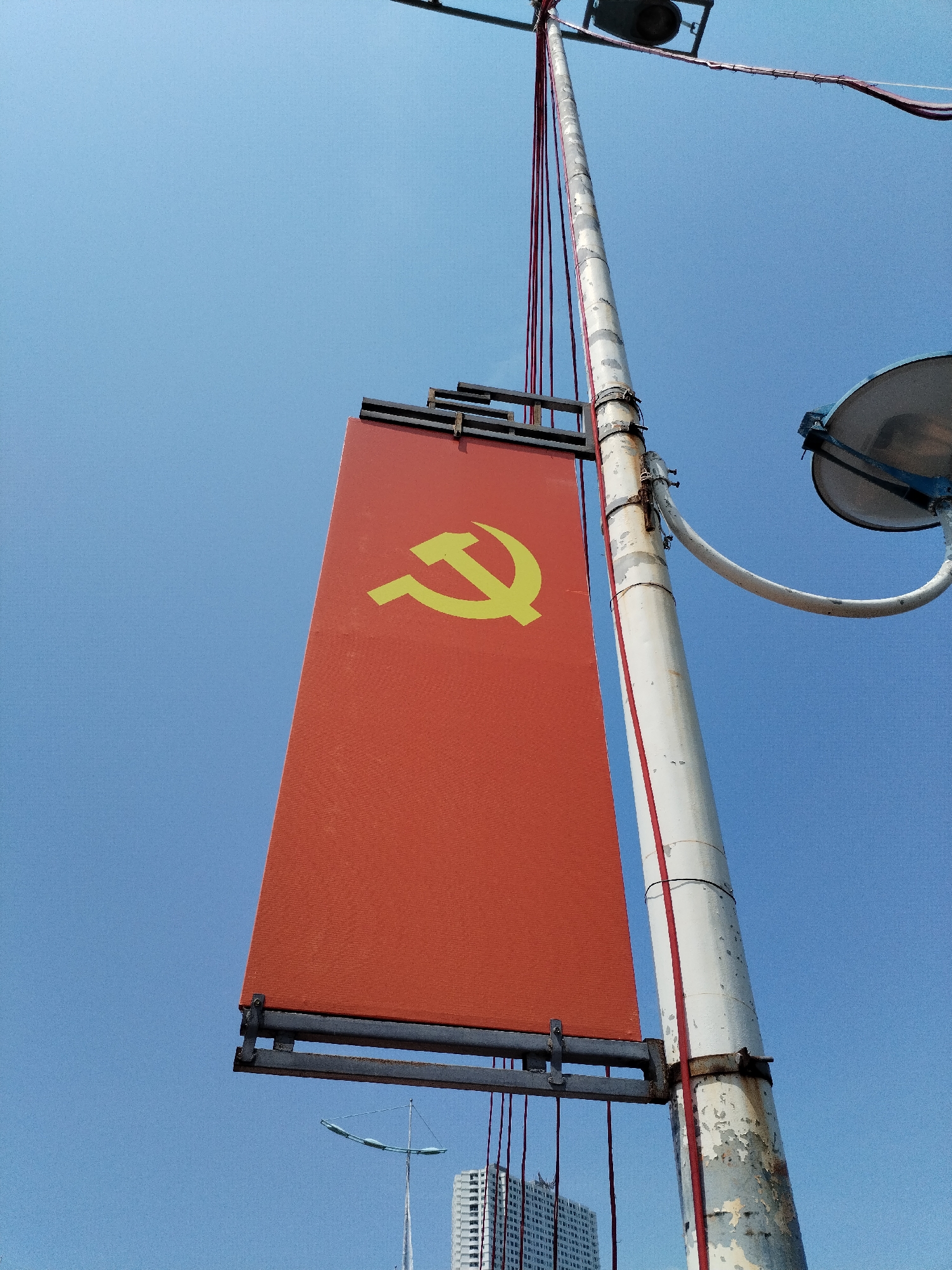 A low-angle shot reveals a weathered, white light pole with a red banner displaying a yellow hammer and sickle symbol attached to it. The sky is a clear, light blue. In the background, buildings are visible, suggesting an urban setting. Based on the provided location, this scene appears to be in Bac Nha Trang Ward, Vietnam.