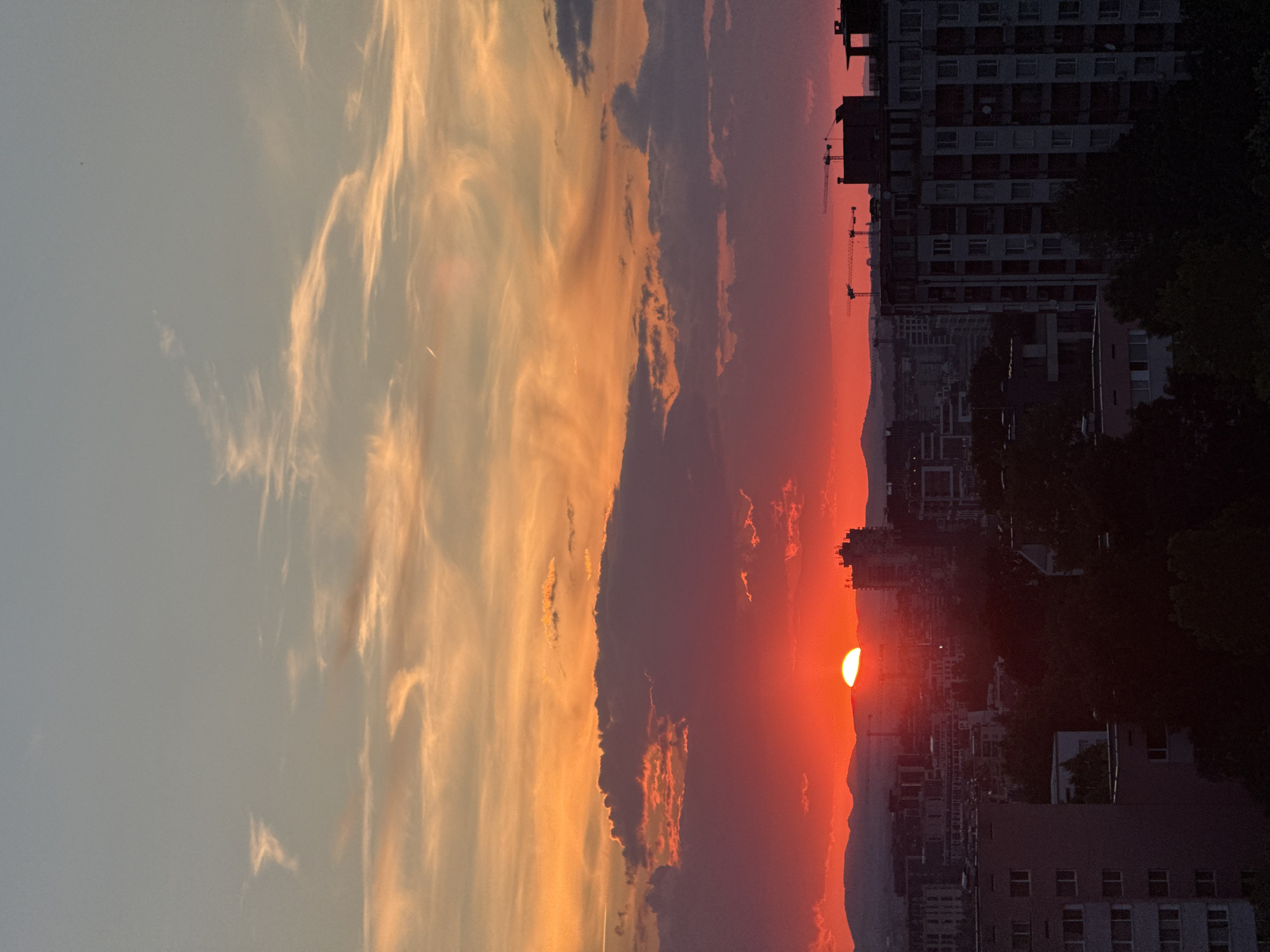 The image depicts a vibrant sunset over the cityscape of Sofia, Bulgaria. The sky is ablaze with a gradient of colors, transitioning from a pale blue at the top to fiery oranges and reds near the horizon. Wispy clouds are scattered across the sky, catching the golden and crimson hues of the setting sun.
Dominating the lower half of the image is a silhouette of the city, characterized by various buildings, including apartment blocks and a few taller structures. Cranes are visible, suggesting ongoing construction. The sun itself is a brilliant, semicircular orb, partially obscured by a dark, horizontal band of clouds and the distant mountain range. The foreground is composed of dark, silhouetted trees and the tops of buildings. The scene captures a moment of natural beauty juxtaposed with an urban environment at dusk.