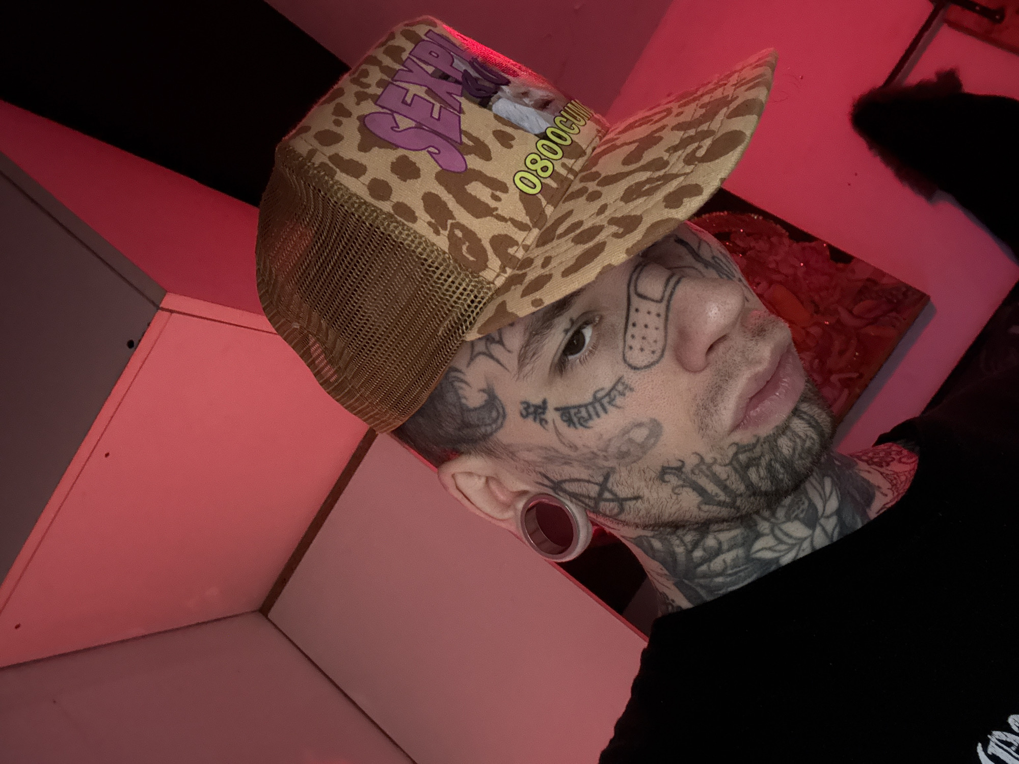 A young adult male, likely posing for a portrait or selfie, is the central subject of the image. He is looking directly at the camera with a neutral to serious expression. His face, neck, and upper chest are extensively covered in tattoos, including a band-aid design on his right cheekbone and the Sanskrit phrase "अहं ब्रह्मास्मि" (Aham Brahmasmi) in Devanagari script on his right temple. He also has large ear gauges in both ears.
He is wearing a brown and tan leopard print trucker hat with a mesh back. The hat prominently displays purple text "SEXY" and yellow text "0800CUM." He is dressed in a black t-shirt, visible on his right shoulder and upper chest.
The background appears to be an indoor setting, strongly illuminated by red lighting that casts a vibrant hue across various geometric surfaces, possibly walls or shelving. A dark, indistinct object is visible in the upper right of the frame, possibly a blurred painting or fabric. The overall lighting creates a dramatic and stylized atmosphere. The image is set in Buenos Aires, Argentina.