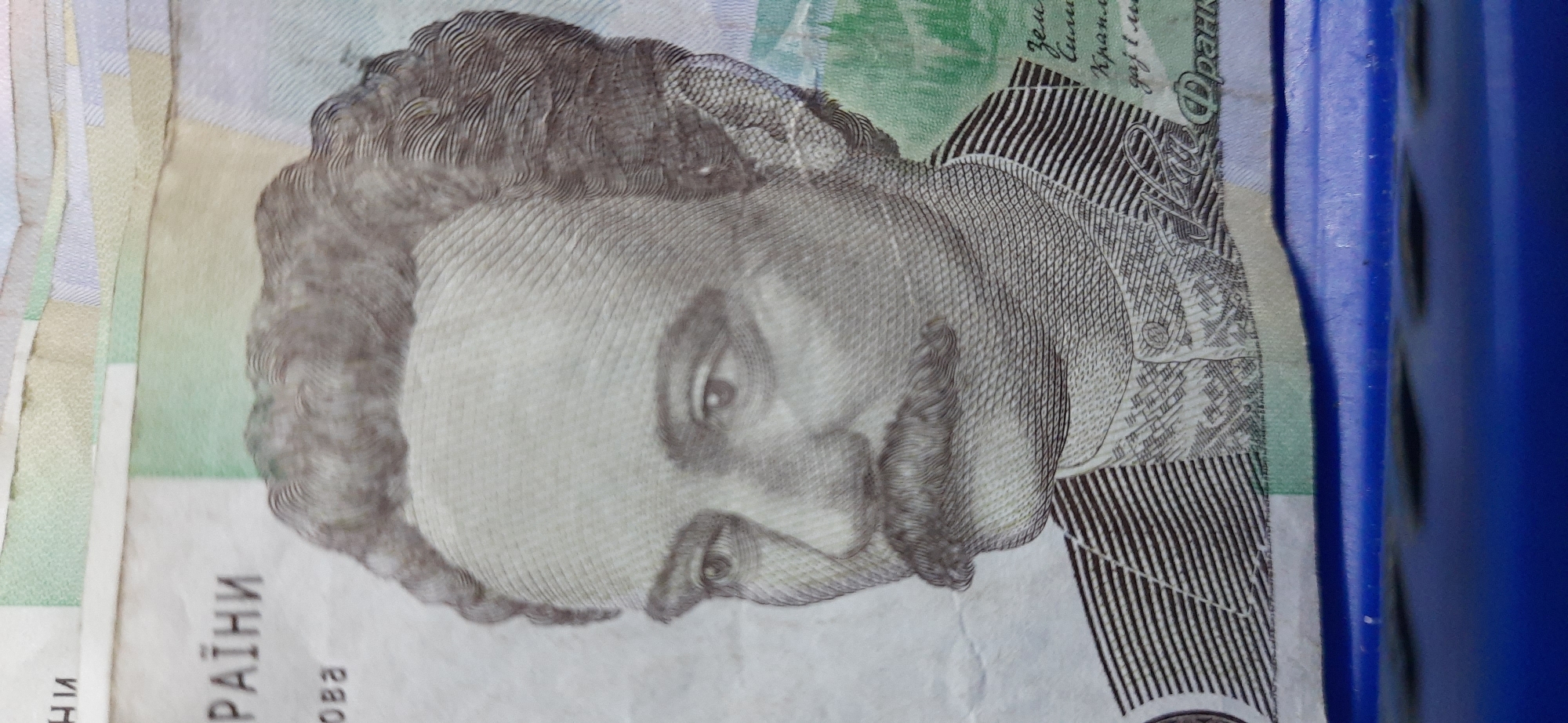 The image shows a close-up of a Ukrainian banknote featuring a portrait of Ivan Franko. The text on the banknote includes "УКРАЇНИ" (Ukraine) and "ГРИВНЯ" (Hryvnia). The background of the banknote displays a landscape scene with mountains and greenery. The bottom portion of the image shows the edge of the banknote resting on a blue, textured surface. There is no indication of the city of Dnipro, Ukraine, or any specific location within it, in this image.