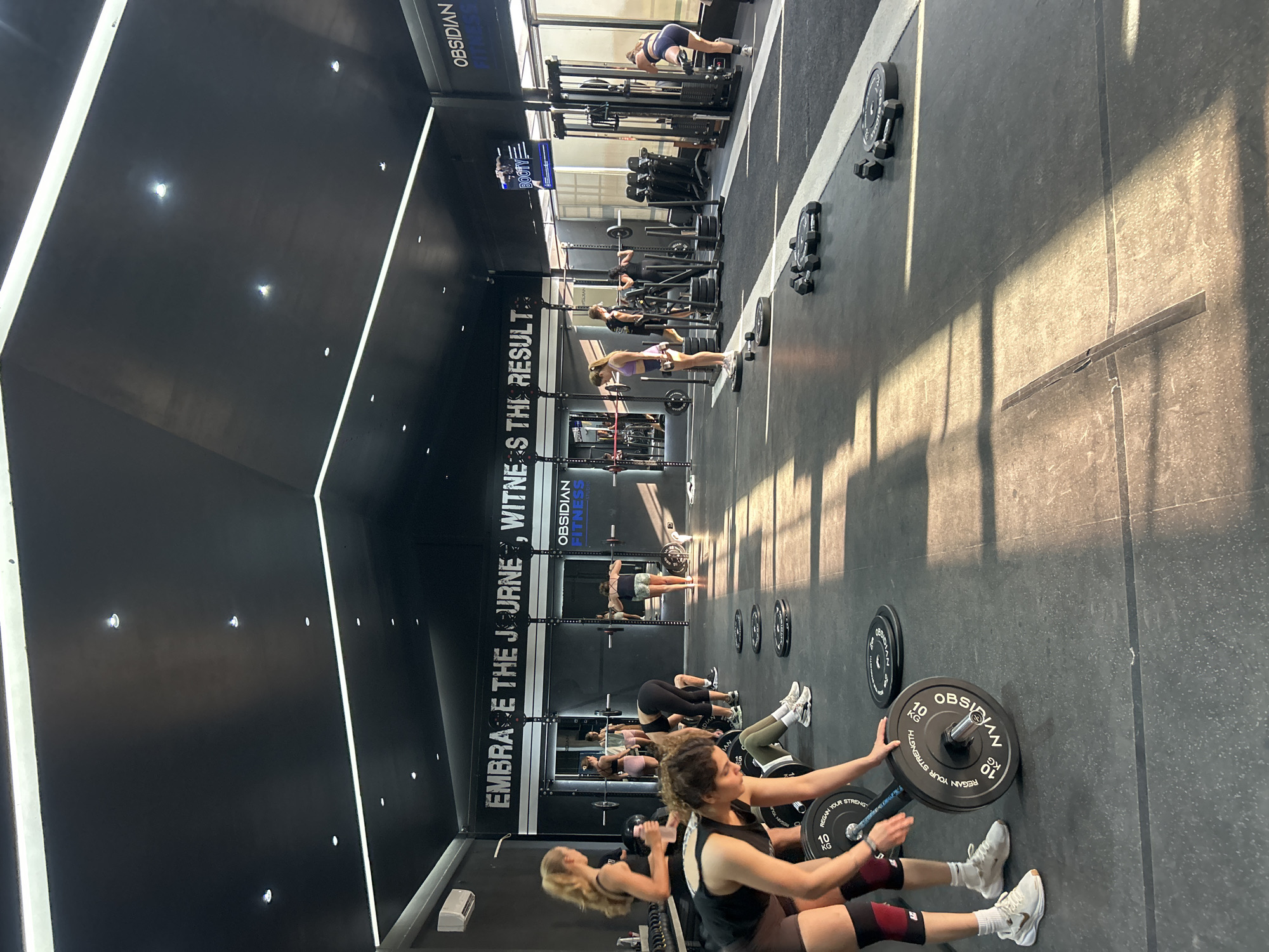 The image depicts a group of women engaged in a workout at a gym. The gym features a modern aesthetic with a dark ceiling and ambient lighting. Several women are actively participating in exercises, with barbells and weight plates visible on the floor. In the foreground, a woman is positioned near a barbell with weight plates. In the background, other women are seen performing various strength training exercises. The gym environment includes weight racks, sleds, and other fitness equipment. A motivational quote, "EMBRACE THE JOURNEY, WITNESS THE RESULT," is displayed on the wall. The location is identified as Obsidian Fitness Studio in Mengwi, Indonesia.