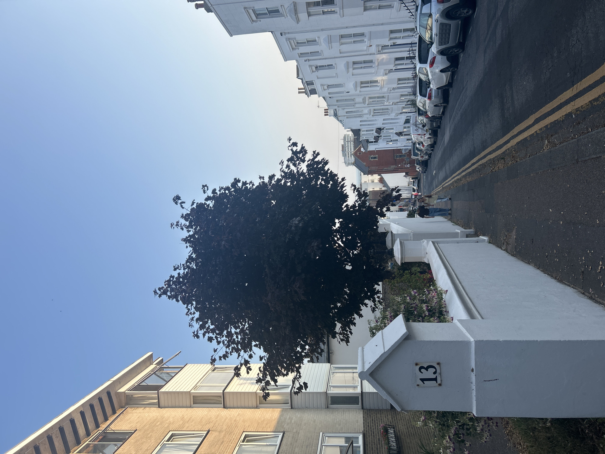 This is a street scene in the city of Brighton, United Kingdom, featuring a row of white terraced houses and parked cars on the right side of the road, and a building with a light-colored facade and balconies on the left. A dark-leaved tree is prominently positioned between the two sides of the street. A white wall with a decorative post displaying the number "13" runs along the left sidewalk. Two individuals, a man and a woman, are walking away from the camera on the left sidewalk. The sky is clear and blue.