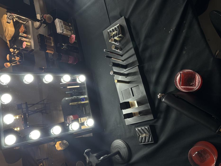 A vanity mirror with illuminated bulbs sits on a black surface with various makeup products. These include brushes, lipsticks, eyeshadow palettes, and foundation. Two drinks with large ice cubes and a black cylinder object also appear on the surface, suggesting a relaxed or professional makeup setting.