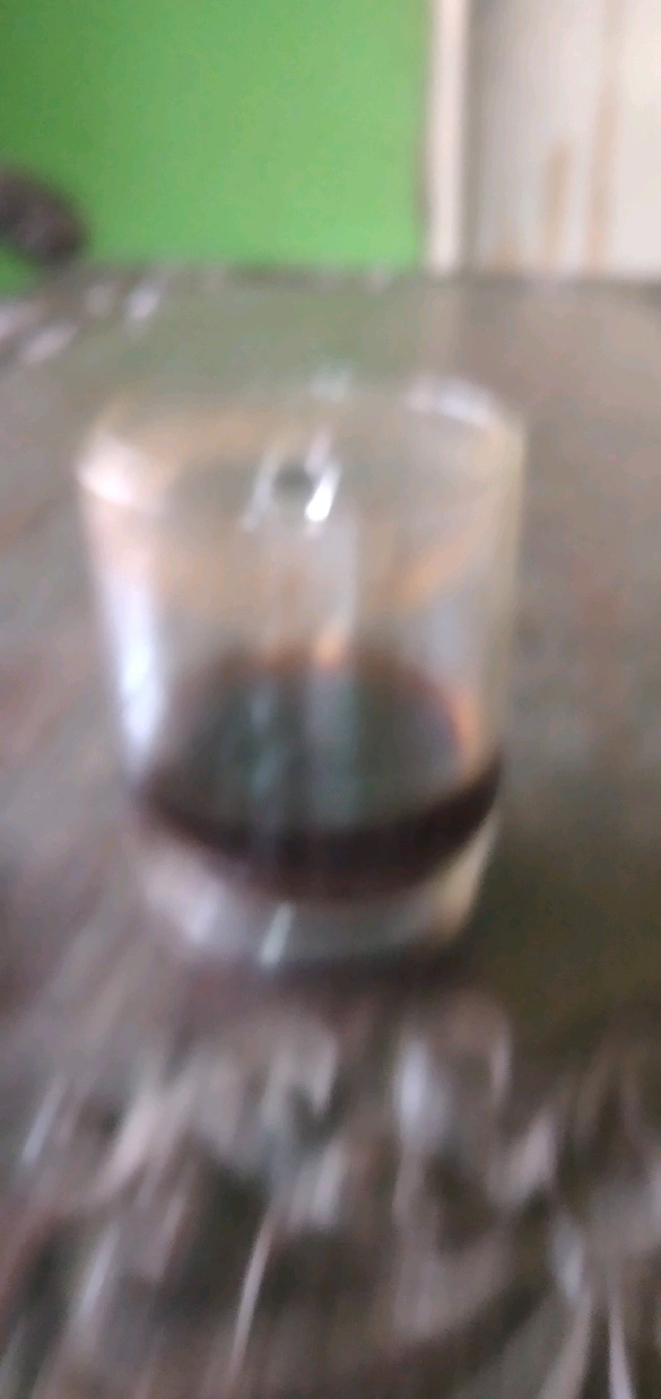 In Rahong, Indonesia, on a blurred metallic surface, a glass is partially filled with a dark liquid. The background is also blurred, with a green wall on the left and a lighter, indistinct wall to the right. The lighting creates reflections on the glass and the surface. No people are visible in the image.
