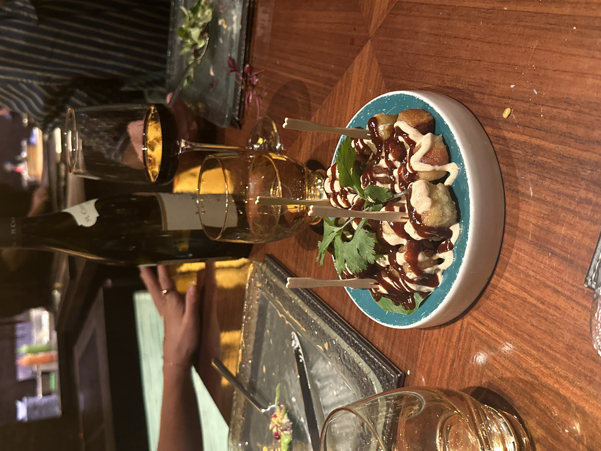 The image captures a dining table scene, likely indoors, with a wooden surface. There's a plate of food featuring small, skewered appetizers drizzled with sauces and garnished with green herbs, possibly cilantro. A half-empty wine bottle and two stemmed glasses, one appearing empty and the other with a golden liquid, suggest a meal or tasting. An arm with a visible ring on a finger reaches across the table, possibly towards a partially eaten dish on a square plate with silverware. Another empty glass sits near the partially eaten dish. The environment is somewhat dimly lit, indicating an evening or subdued setting. Based on the sophisticated ambiance, the location is possibly a restaurant within the city of London, United Kingdom.