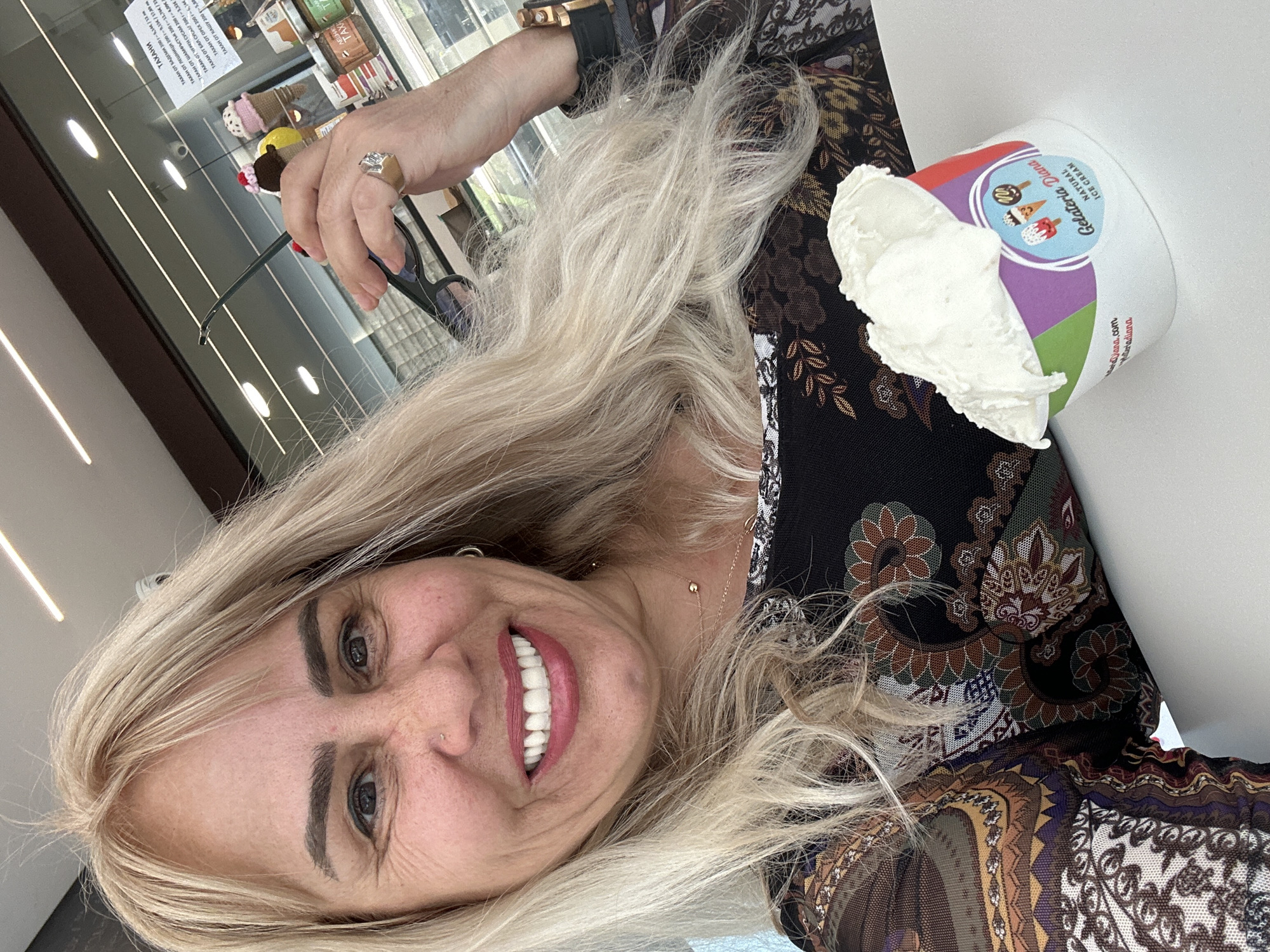 A smiling woman with long, flowing blonde hair takes a selfie indoors. She is positioned slightly off-center, looking directly at the camera with a wide, joyful smile that reveals her teeth. Her dark eyebrows and eye makeup are prominent, and she has a small nose piercing. She wears a dark top adorned with an intricate, colorful paisley and floral pattern in shades of brown, gold, green, and blue.
On her right wrist, she wears a gold-colored watch with a black strap, and a ring with a large stone is visible on her finger. Her left hand, partially visible, holds a pair of dark-framed glasses.
Beside her, on a white surface, is a white cup of "Gelateria Diana NATURAL ICE CREAM," from which a large scoop of white ice cream overflows. The cup's label features a purple and green design with cartoon ice cream cones, and the website "diana.com" and the word "radiona" are printed on it.
The background shows a reflective surface, likely a display case, with various items and bright interior lighting. A sign in Bulgarian is visible in the reflection, detailing different types of tahini (from almonds, hazelnuts, pistachios, black sesame, white sesame, walnuts, and cashews) along with their weights (200g) and prices in both Euros and Bulgarian Leva. This suggests the scene is an ice cream parlor or a sweet shop, consistent with the location hint of Sofia, Bulgaria. The atmosphere appears happy and relaxed.