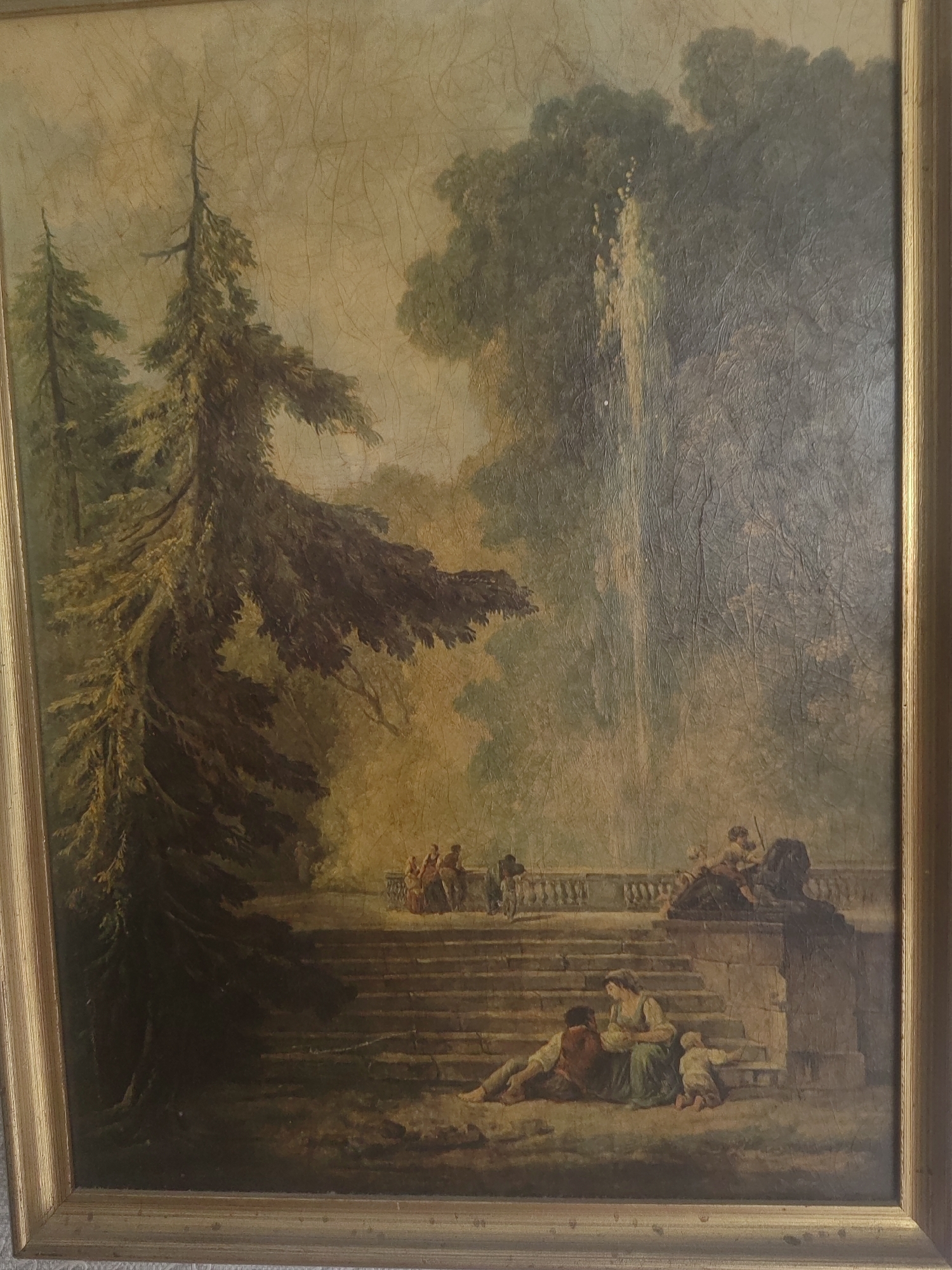 The painting depicts a pastoral scene with a large evergreen tree dominating the left foreground, its branches reaching across the upper half of the canvas. In the middle ground, a series of stone steps leads up to a terrace with a balustrade. A fountain is visible on the right side of the terrace, with water spraying upwards. Several figures are present: a couple with a child are seated on the lower steps, with the woman holding an infant. Other figures are seen on the terrace, some standing and others walking. The environment appears to be a park or garden, with lush greenery and a soft, diffused light. There is no discernible indication of the city of Izhevsk, Russia, or any specific urban location. The painting is framed in a gold-colored wooden frame.