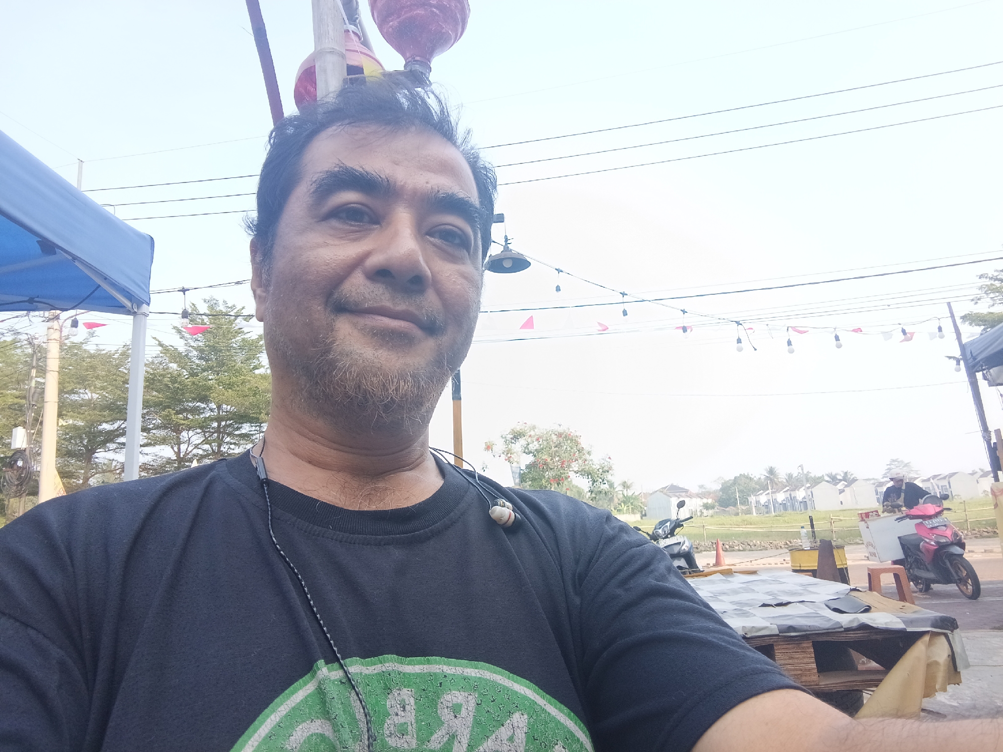 A middle-aged man with dark hair and stubble, wearing a black t-shirt with a green graphic, is taking a selfie outdoors. He has earphones around his neck. The background shows a bright, overcast sky with power lines and some decorative lights strung between poles. To the left, a portion of a blue tent is visible. Further in the background, there are trees, a residential area with white houses, and a road with motorcycles. One person is on a pink and black motorcycle, and another motorcycle is parked. There are also some cones and what appears to be a market stall with a checkered tablecloth. The overall setting suggests a market or street-side location in Ciseeng, Indonesia.