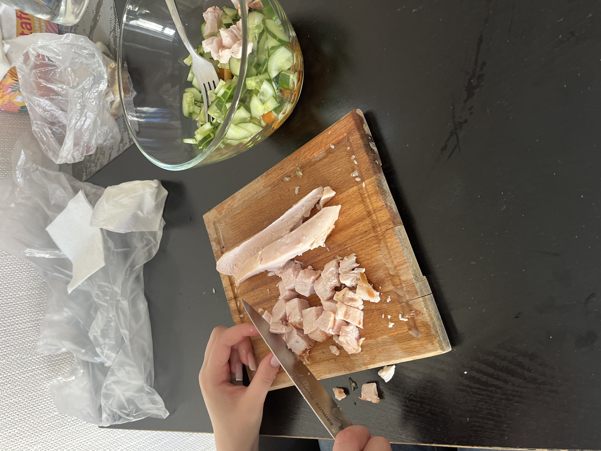 The image shows a close-up, high-angle view of a person's hands chopping cooked chicken breast on a wooden cutting board. The chicken is being diced into small, bite-sized pieces. The cutting board is placed on a dark, reflective surface, likely a table. To the right of the cutting board, a glass bowl contains a salad made with chopped cucumbers and what appears to be pieces of ham or more chicken. A fork rests in the salad bowl. In the background, there are various items, including a crumpled plastic bag, paper napkins, and a bottle of water. The overall impression is that of food preparation in a home kitchen. The image does not contain any elements that would allow for the determination of its specific location within the city of Perm, Russia, or any other city.