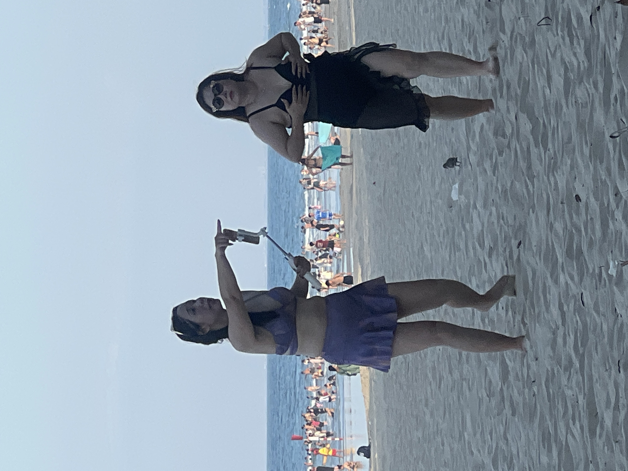 Two women are standing on a sandy beach, with the ocean and a crowded shoreline in the background. The woman on the left, facing away from the viewer, is wearing a purple bikini with a ruffled skirt and is holding a selfie stick with a phone. She has her left arm extended, pointing towards the right. The woman on the right, facing the viewer, is wearing a black bikini with a sheer black skirt and sunglasses. She has her hands on her chest. In the background, many people are visible in the water and on the shore, suggesting a popular beach. The overall setting is a daytime scene at a beach in Đà Nẵng, Vietnam.