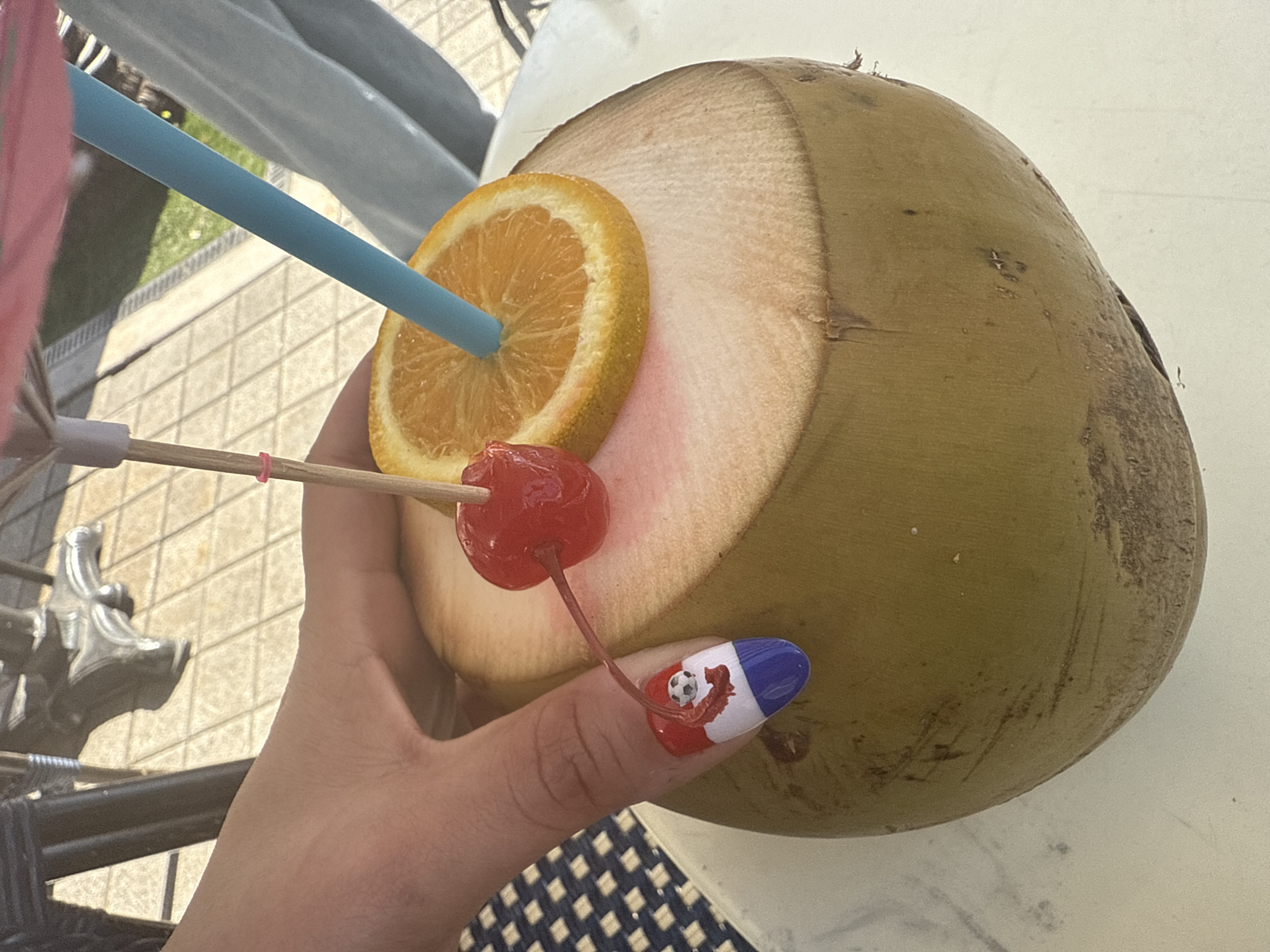 In Miami Beach, United States of America, a person's hand with a red, white, and blue nail design featuring a small soccer ball is holding a fresh coconut. The coconut has been opened and is presented as a drink, garnished with a slice of orange, a maraschino cherry on a cocktail pick, a small pink umbrella, and a blue straw. The scene appears to be outdoors, with a glimpse of a tiled patio and outdoor furniture in the background, suggesting a casual, possibly resort-like setting.