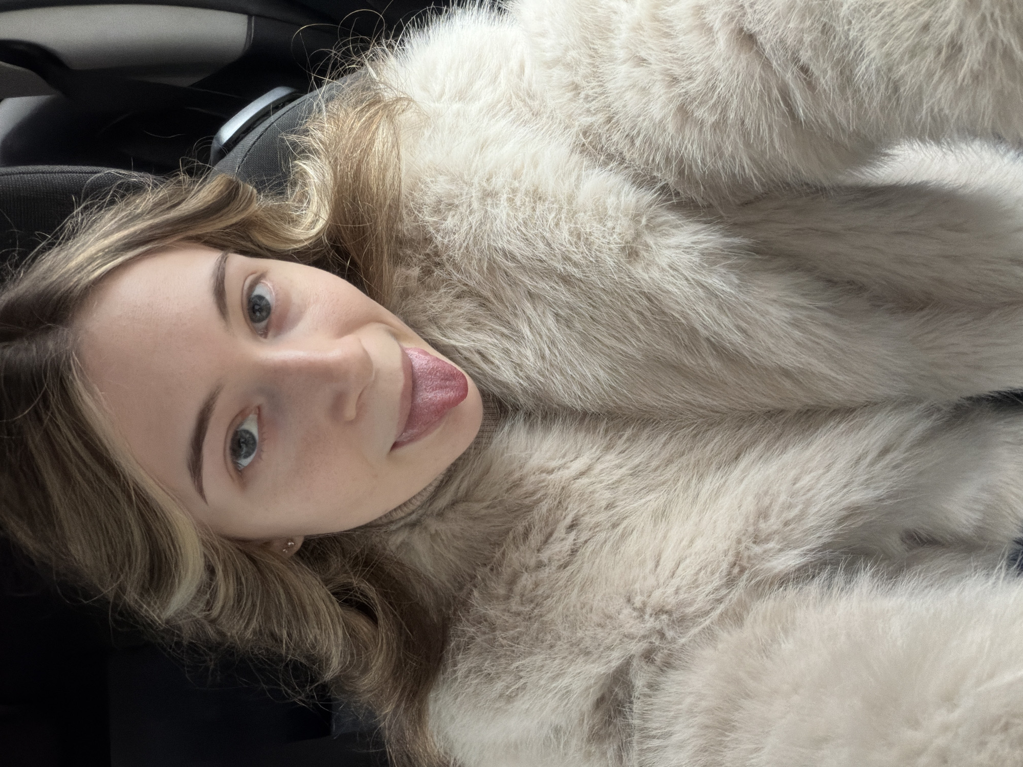 A young woman with fair skin, light brown hair with blonde highlights, and blue eyes is visible. Her tongue is slightly protruding. She is wearing a white fur coat. The background indicates she is likely in a car. It is not possible to determine her location as being in Plovdiv, Bulgaria from the information provided.