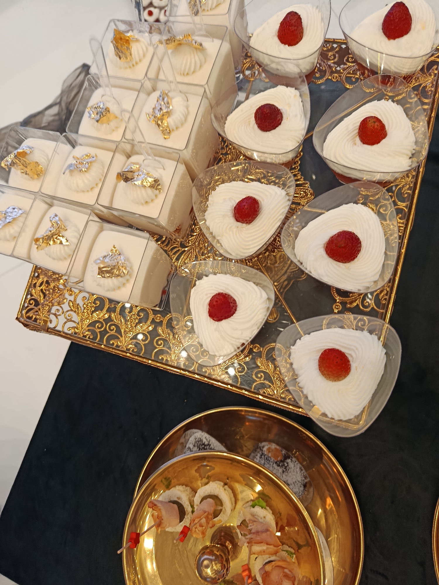 A close-up, high-angle shot shows a spread of desserts on a gold tray. The desserts are presented in small, clear plastic cups and are arranged in rows. The first row, closest to the viewer, features small rolls with red and white filling, speared with red toothpicks, on a gold plate. Behind these, a larger gold plate holds what appear to be small chocolate cakes dusted with powdered sugar. The main focus of the image is a gold tray holding numerous small desserts. Some of these are layered white desserts topped with gold leaf and served with small plastic spoons. Others are white whipped cream desserts topped with a single strawberry, presented in heart-shaped clear plastic containers. The tray itself has an ornate gold design along its edges. The background is a dark, textured fabric.