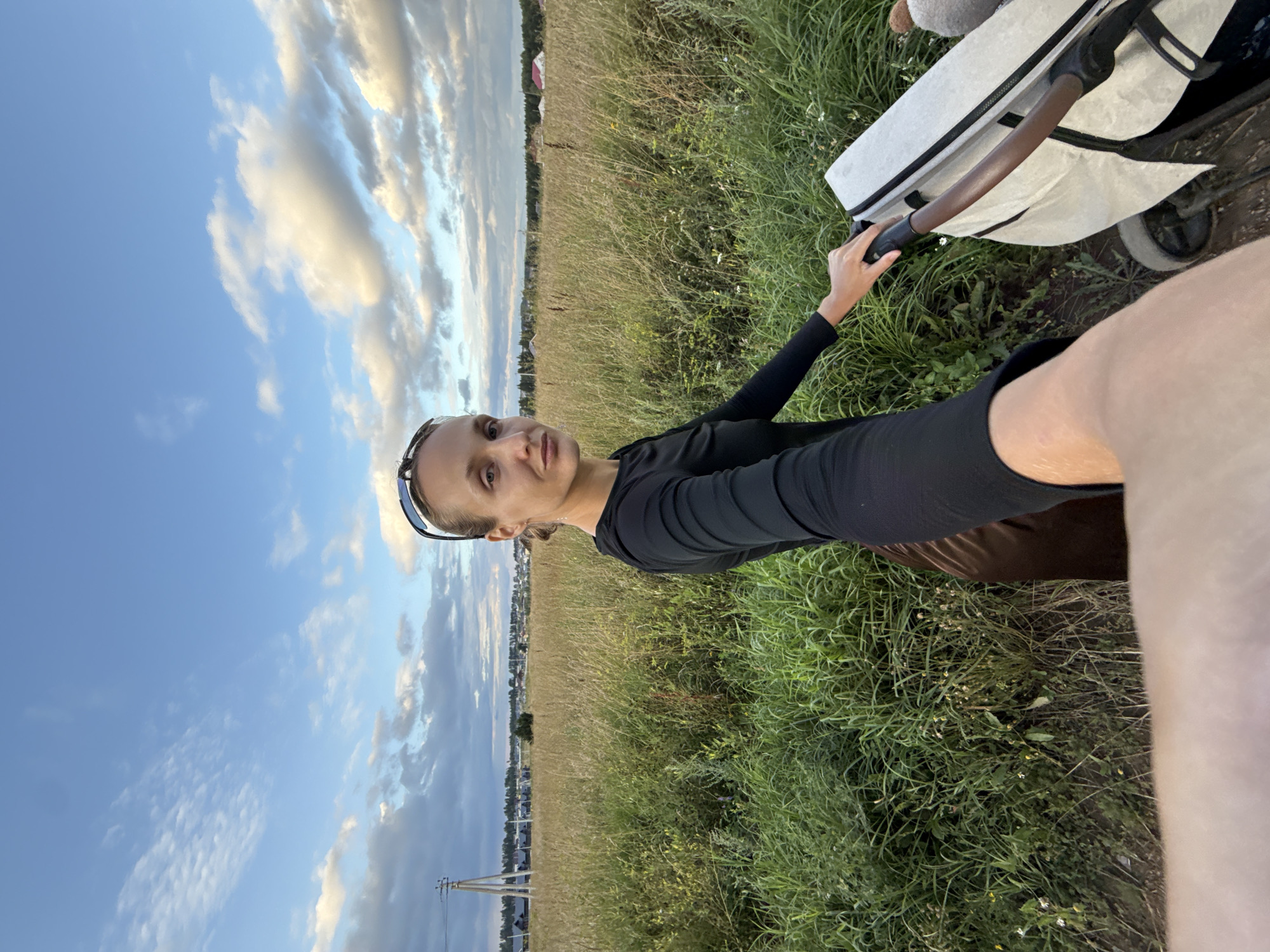 A medium shot captures a woman holding the handle of a white stroller while walking through a field of tall grass and wildflowers under a cloudy sky. The woman, with blonde hair pulled back and wearing sunglasses on her head and a black long-sleeved shirt, looks directly at the camera. Her brown skirt is visible. In the background, a village with houses and a power line is nestled amongst trees. The scene appears to be in or near the village of Podshivalovo, Russia. The lighting suggests it is late afternoon or early evening.