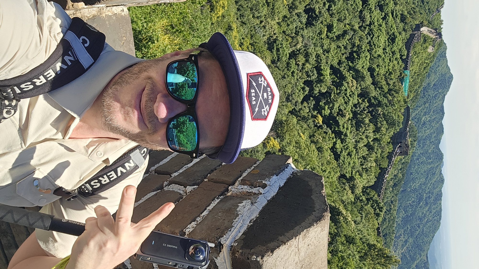 A man wearing a white and blue cap, sunglasses, and a light-colored shirt is taking a selfie on the Great Wall in Huairou District, China. He is smiling and making a peace sign with his right hand. He is holding a black Insta360 X3 camera. The Great Wall stretches across a lush, green, mountainous landscape in the background. The sky is bright and clear.