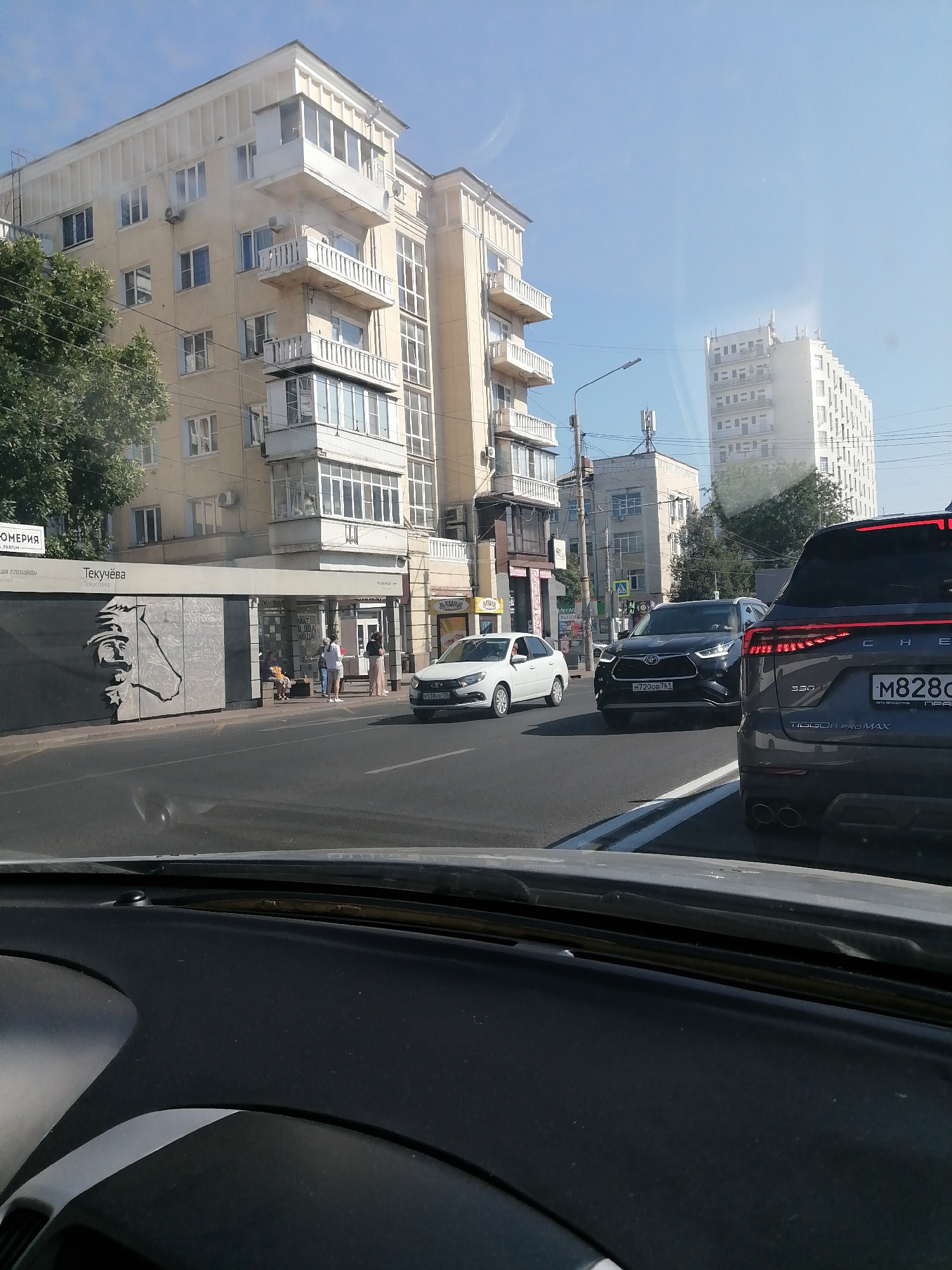 A street scene in Rostov-on-Don, Russia, captured from inside a vehicle. The view includes a modern apartment building with balconies, a bus stop with a metal relief sculpture, several vehicles on the road, and other buildings in the background. The sky is clear and blue, indicating a sunny day. The perspective suggests the vehicle is in traffic, with other cars ahead and to the side. The bus stop has the name "Текучёва" (Tekucheva) visible. Several people are waiting at the bus stop or walking nearby.