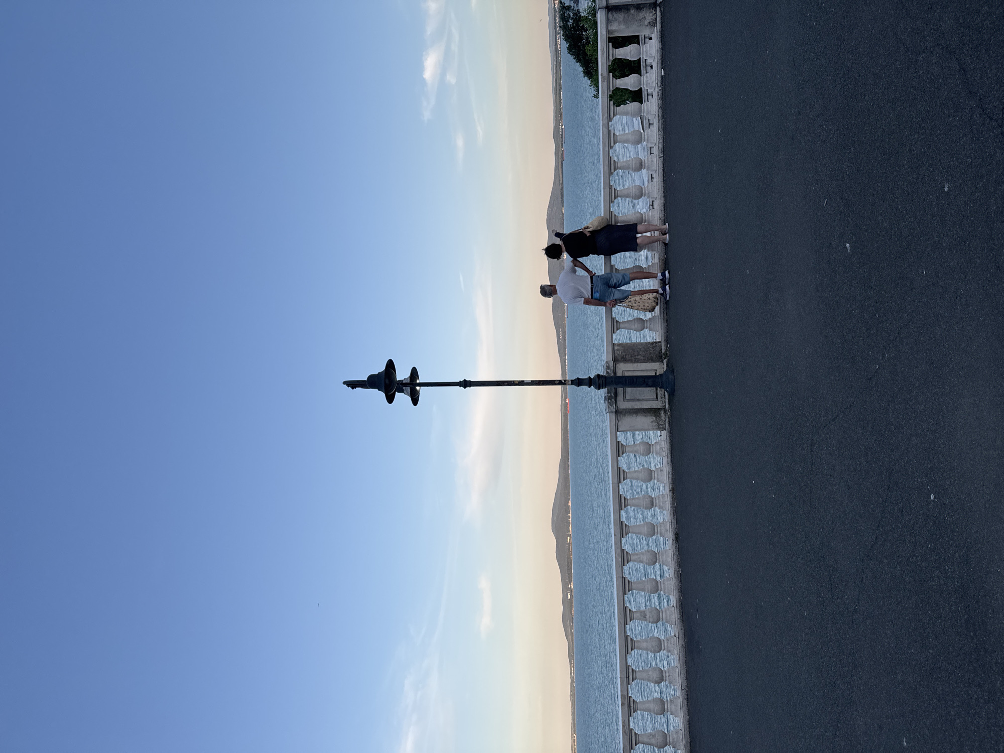 The image depicts a serene view of the Burgas Bay in Burgas, Bulgaria. In the foreground, a couple stands near a decorative balustrade, gazing out at the calm water. The man, wearing a white t-shirt and denim shorts, holds a patterned bag. The woman, in a dark skirt and top, points towards the horizon. A lamppost stands to their left, a silhouetted sentinel against the expansive blue sky. In the distance, rolling hills form a picturesque backdrop, with a few boats visible on the water. The scene is bathed in the soft light of what appears to be late afternoon or early evening, contributing to the tranquil atmosphere of the coastal promenade.