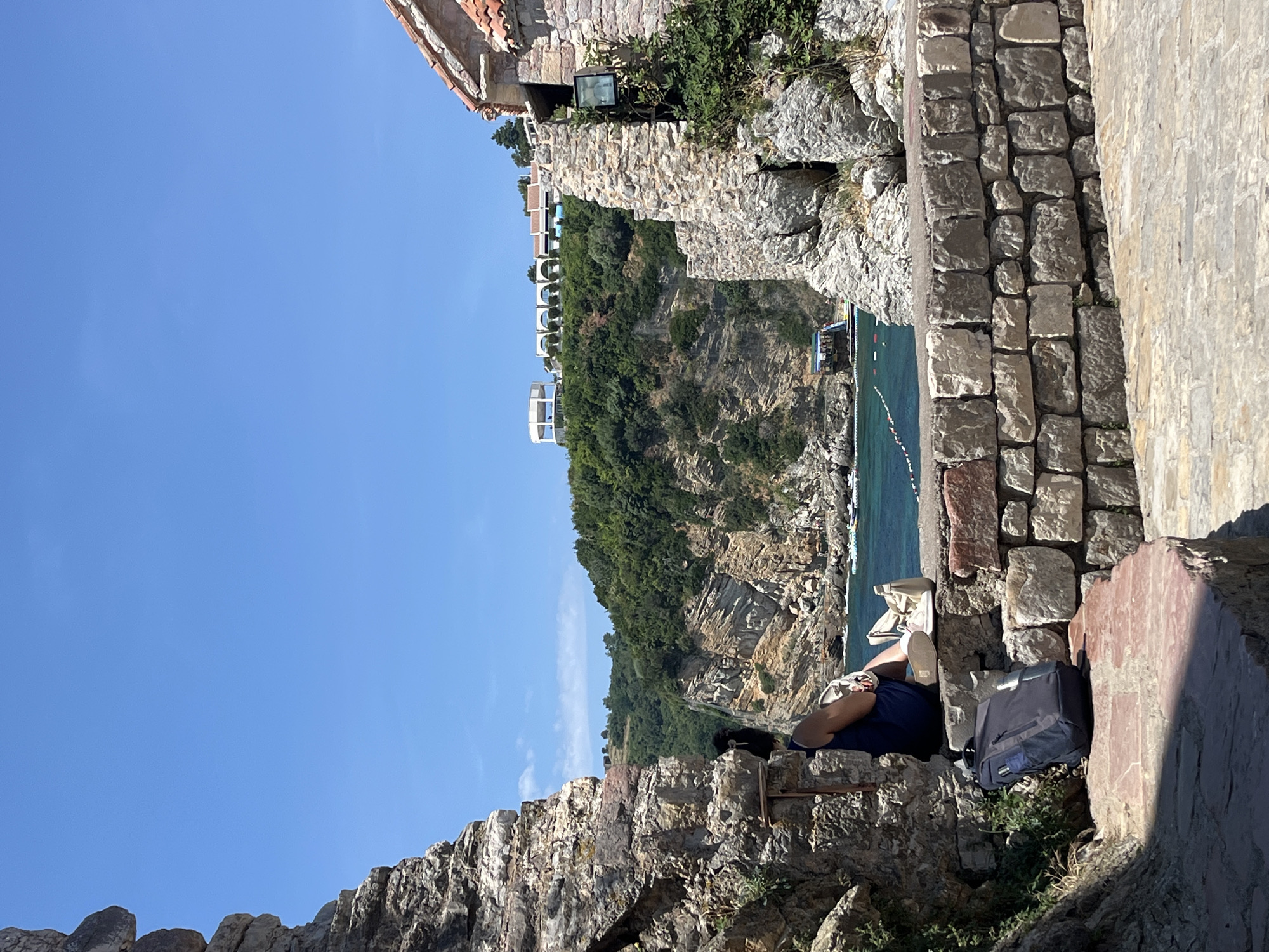 In Budva, Montenegro, a person is seated on a stone wall, facing away from the camera, looking out at a bay. The person is wearing a dark blue dress and has a light-colored bag and shoes next to them. A black backpack rests on the ground in front of them. The stone wall is constructed from irregular grey and tan stones. Beyond the wall, the turquoise water of the bay is visible, with a rope and buoys marking an area. Steep, rocky cliffs covered in green vegetation rise from the water, and buildings with red-tiled roofs are visible on the hilltop in the distance. The sky is a clear, bright blue. The foreground is a textured, pinkish stone pathway. The scene is framed by rough, grey stone structures on either side.