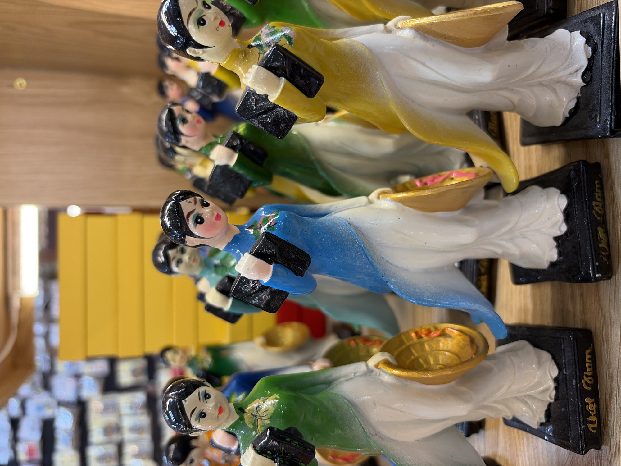 This is a close-up, eye-level, slightly tilted-down shot of a row of small, decorative figurines displayed on a wooden surface, likely a shelf or table. The figurines are of women dressed in traditional Vietnamese áo dài. Each figurine has a black bobbed hairstyle, a light skin tone, rosy cheeks, and red lips. They are all holding a black rectangular object in their hands. The figurines are standing on black rectangular bases, some of which have "Việt Nam" inscribed on them in gold lettering.
The figurines are arranged in multiple rows, with the front row being the most in focus. From left to right in the front row, the first figurine is wearing a green áo dài and holding a golden conical hat in her left hand. The next figurine is wearing a blue áo dài and holding a golden conical hat in her right hand. The third figurine, partially visible, is wearing a yellow áo dài. Further back, other figurines in green, blue, and yellow áo dài can be seen.
The background is blurred but suggests a store or market setting. Behind the figurines, there are stacked yellow boxes. In the far background, out-of-focus shelves with what appear to be more products or displays are visible. The lighting appears to be ambient, possibly from overhead lights, creating some highlights on the glossy surfaces of the figurines. The overall impression is a display of souvenir items. No specific location in Đà Nẵng, Vietnam can be identified from the image itself.