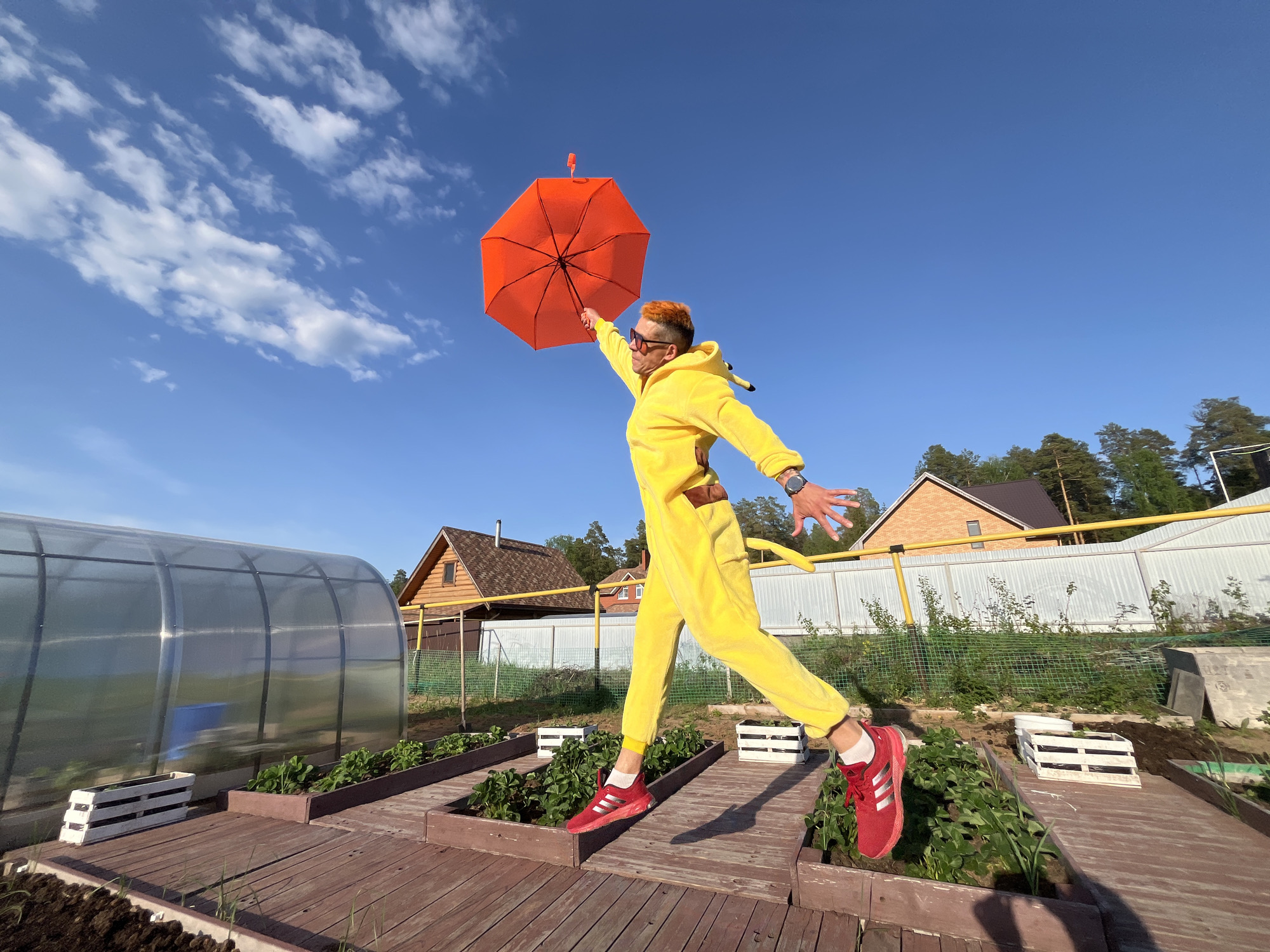 A person wearing a yellow Pikachu jumpsuit and red Adidas sneakers is jumping in the air, holding an orange umbrella, on a sunny day in what appears to be a backyard garden. The person has orange hair and is wearing sunglasses. The garden features wooden planters, a greenhouse, and a white fence in the background. Houses and trees are visible behind the fence. The location is likely the city of Chaikovsky, Russia.