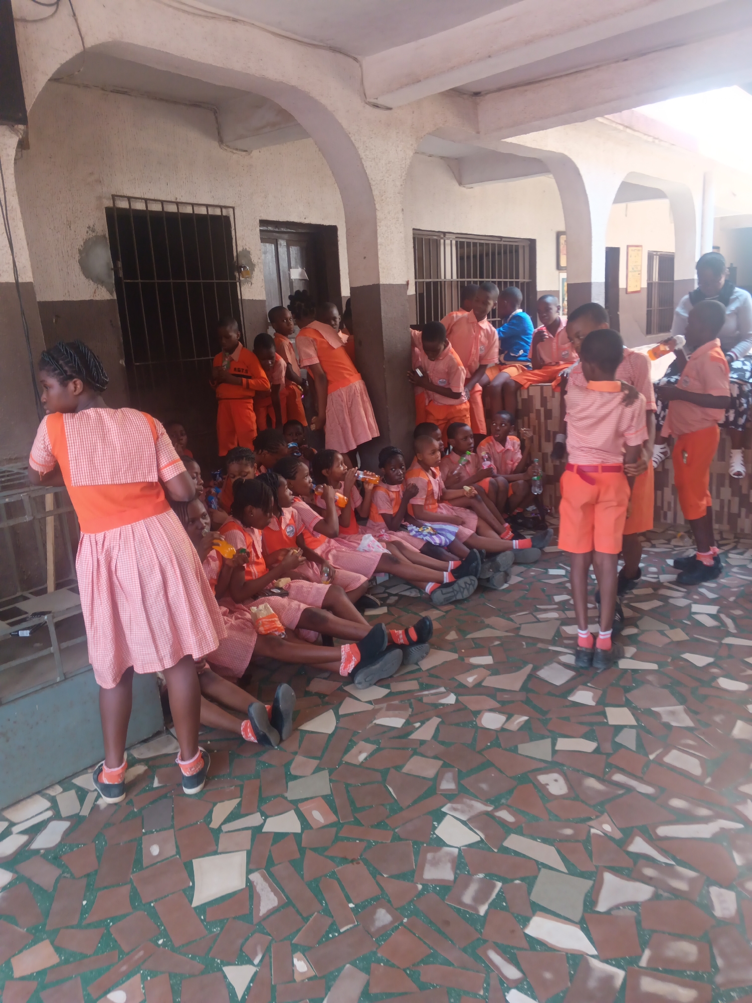 In Onitsha, Nigeria, a group of children, likely students, are gathered in an outdoor covered area with a tiled floor. The children are predominantly wearing orange and white school uniforms. Some are seated on the ground or on a low ledge, while others are standing. Many of the children are holding or drinking from bottles, suggesting a break or a refreshment period. The setting appears to be a schoolyard or an outdoor common area of an educational institution, characterized by a textured wall, arched walkways, and barred windows. The time of day seems to be daytime, with sunlight casting shadows, indicating possibly a warm climate. The overall atmosphere appears relaxed and casual, with the children engaged in conversation or simply resting.