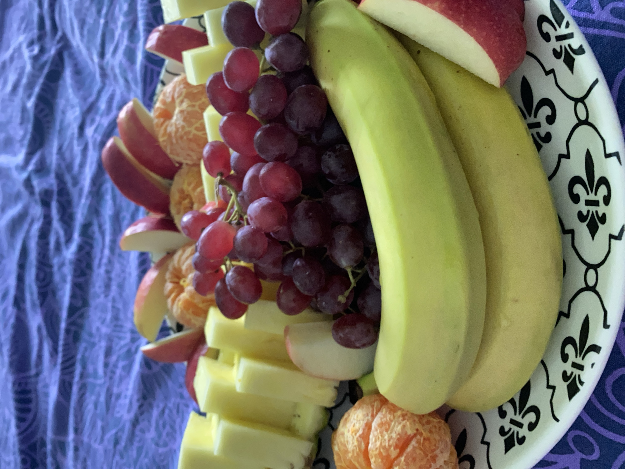A close-up, eye-level shot shows a fruit platter arranged on a white plate with a black fleur-de-lis pattern around the edge. The plate rests on a blue and purple patterned tablecloth. The platter contains banana, red grapes, apple slices, mandarin orange segments, and pineapple chunks. The lighting appears to be natural and indirect. There is no indication of this being in Chaikovsky, Russia. There are no people depicted in the image.