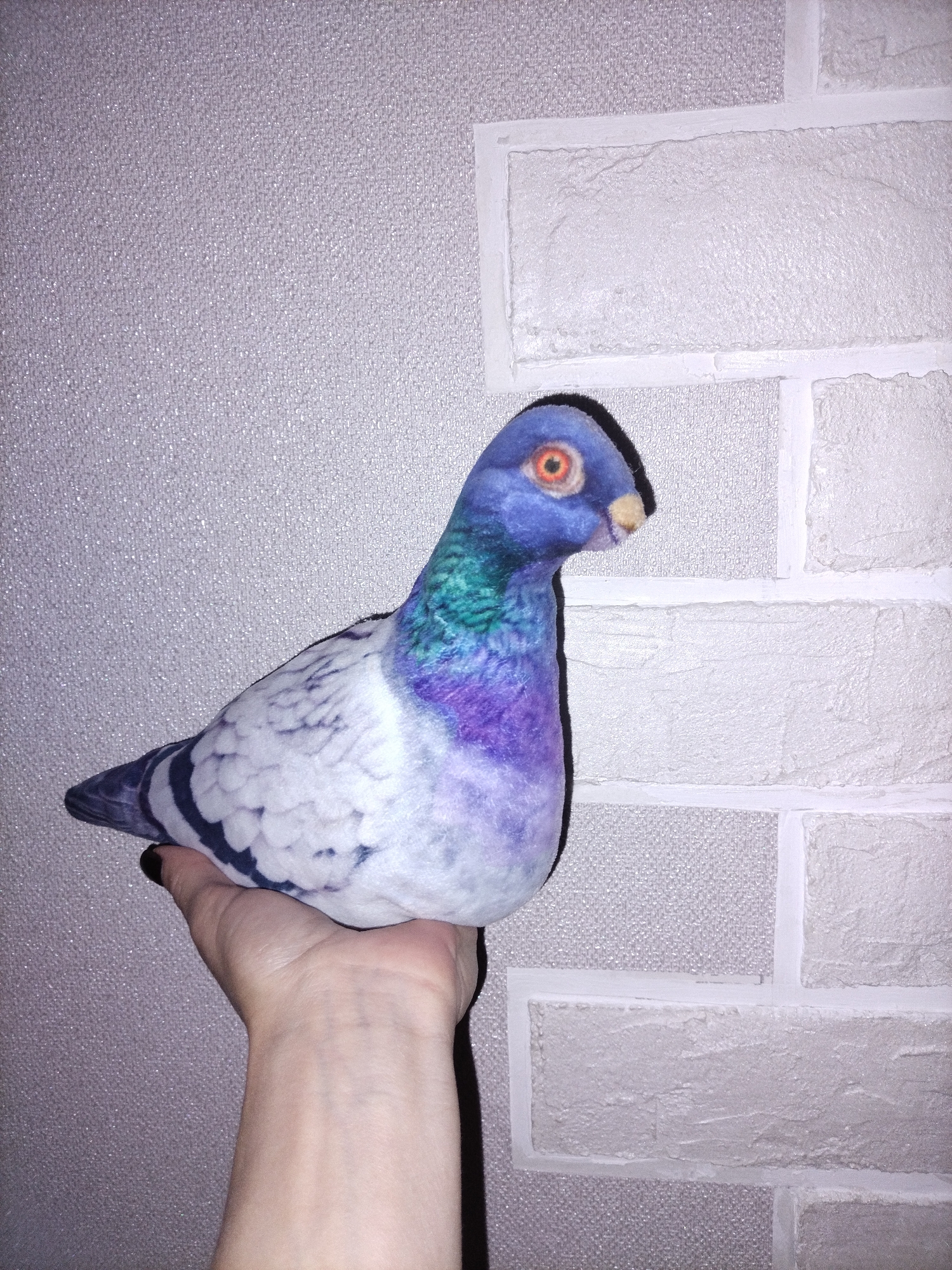 A close-up indoor shot features a human hand holding a plush pigeon against a textured wall. The hand, with a light skin tone and black nail polish on what appears to be the thumb, supports the pigeon from underneath.
The plush pigeon is multicolored, primarily blue and purple on its head and neck, transitioning to white and grey with black striped patterns on its body and wings. Its head has a distinct blue hue, with a prominent orange eye and a black pupil, and a small yellowish beak. The pigeon is positioned to face right, with its profile clearly visible.
The background consists of a wall covered with two different types of wallpaper. On the left, there's a light grey or silver wallpaper with a fine, granular, and slightly sparkling texture. On the right, the wallpaper mimics white brickwork, featuring raised, irregular brick shapes and clear, light-colored grout lines. The junction between the two wallpaper types is visible, running vertically down the image.
The lighting appears artificial, creating a dark shadow of the pigeon and the hand on the wall behind them. No text is visible in the image, and no specific location or time of day can be determined beyond being indoors.