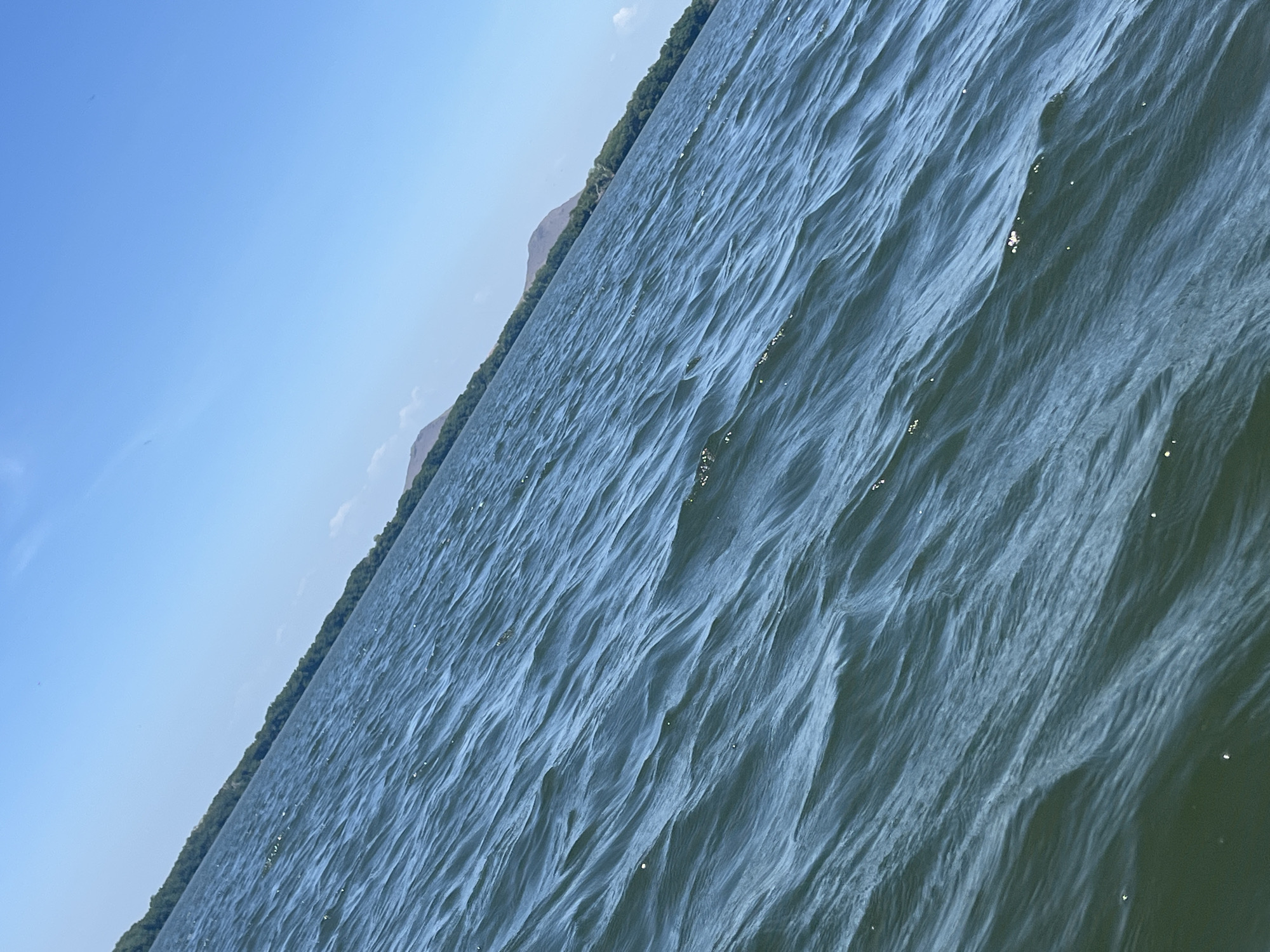 The image shows a body of water, possibly the Laguna de la Restinga in Venezuela, based on the presence of a narrow strip of land with vegetation and hills in the distance. The water has a choppy surface with visible wave patterns and light reflecting off the ripples. The sky above is blue with some wispy clouds.