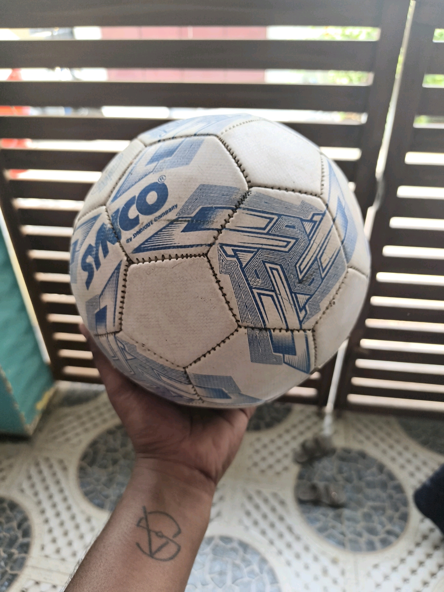A soccer ball is held in a person's hand, positioned in the foreground of the image. The soccer ball is white with blue patterns and features the logo "SYNCO". The background consists of a wooden slatted fence, through which glimpses of greenery and light can be seen. The floor beneath the person is tiled with a circular pattern. A tattoo is visible on the person's wrist. The scene appears to be outdoors, possibly in a residential area or a public space in Coimbatore, India, given the context of the request.
