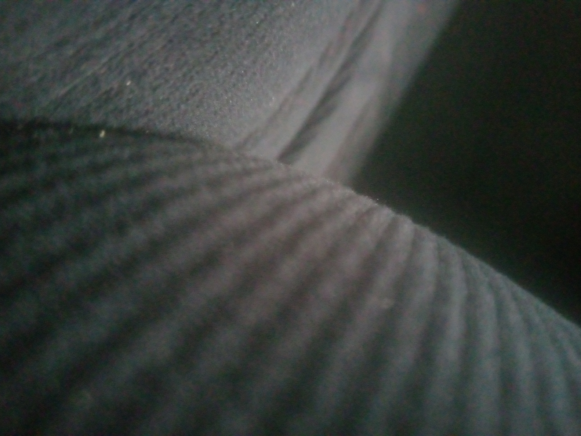 The image is a close-up, out-of-focus shot of a dark, textured fabric, likely upholstery or clothing. The fabric appears to have a ribbed or corded pattern running horizontally across the frame. There are subtle variations in light and shadow, suggesting a curved surface and possibly some folds or creases. The lighting is dim, creating a moody and somewhat indistinct atmosphere. No people, specific objects, or distinct environmental features are clearly visible due to the extreme close-up and lack of focus. The overall impression is of a detail of a larger, unseen object. Given the location context of Jalingo, Nigeria, it is possible this fabric is part of furniture or clothing found in that region, but there are no direct visual cues to confirm this. There is no visible text in the image.
