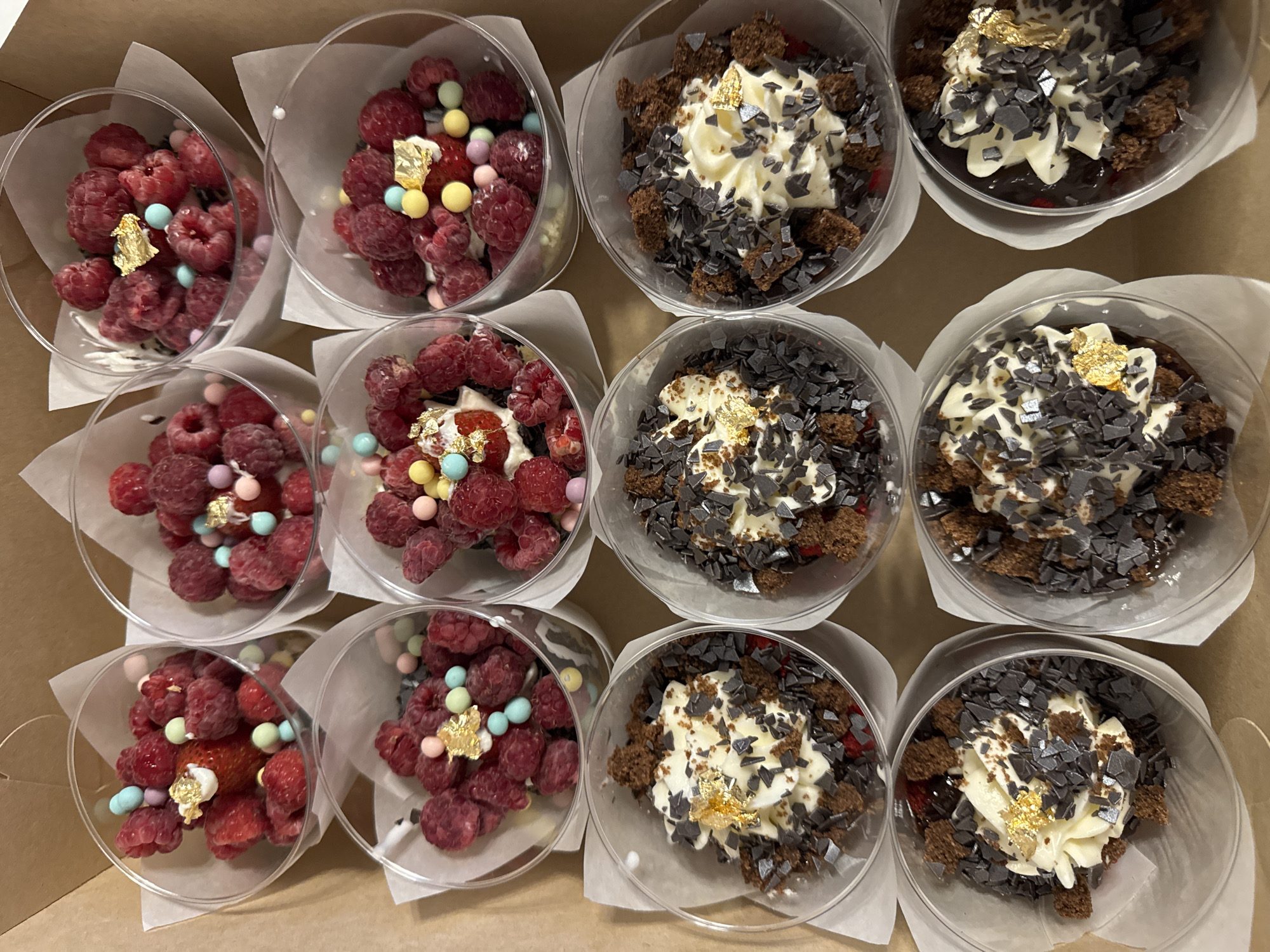 The image shows a top-down view of several individual dessert cups arranged in a cardboard box. There are two rows of desserts, with four cups in the top row and four cups in the bottom row, though the bottom row is partially obscured.
The desserts in the top row appear to be fruit-based, with a generous amount of fresh raspberries filling the clear plastic cups. Some of these cups also contain what appear to be small, colorful candies and pieces of edible gold leaf. One cup clearly shows a whole strawberry.
The desserts in the bottom row are different. They feature a base of crumbled chocolate cake, topped with whipped cream or frosting, and generously sprinkled with dark chocolate shavings. These desserts also have small pieces of edible gold leaf. Some red fruit is visible beneath the cream in these cups.
The environment is indoors, suggested by the cardboard box and the lighting. The location of Razdolnaya, Russia is irrelevant to the visual content of the image.
