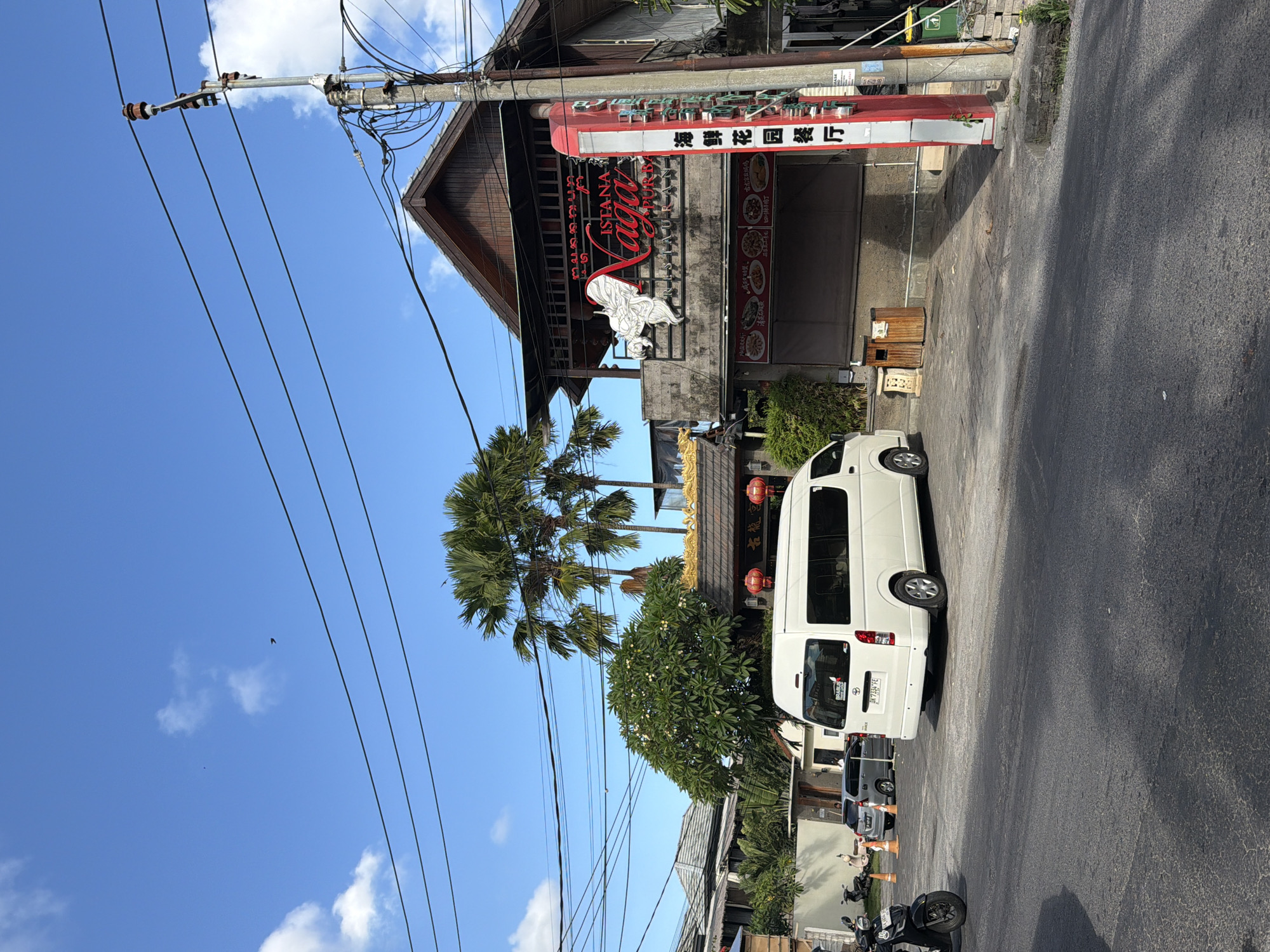 In the city of Kuta, Indonesia, a white van is parked on an asphalt road in front of a two-story building. The building has a sign that reads "Istana Naga Purba Restaurant." There are palm trees and a large green tree with white flowers in front of the building. Overhead, power lines crisscross the bright blue sky, which is dotted with white clouds. To the left of the building, there is a black motorcycle parked. The scene appears to be a commercial area with a mix of residential and business structures.