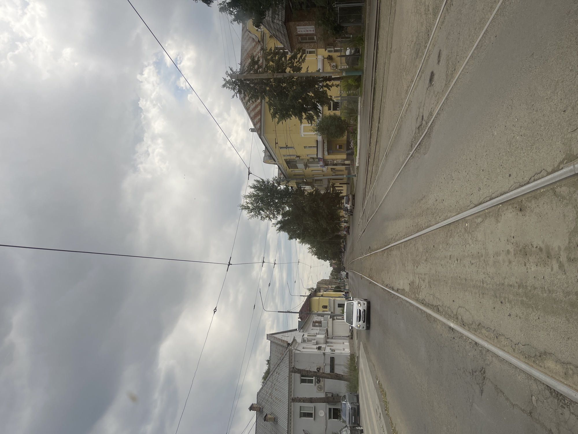The image shows a street in Oskemen, Kazakhstan, with tram tracks running down the center. The sky is cloudy and overcast. On the left side of the street, there are several buildings, including a light-colored building with a grey roof and a yellow building with a red roof. There are also trees and some parked cars. On the right side of the street, there are more buildings, including a large yellow building and a smaller brick building. The tram tracks are laid on a paved surface, and the road is also paved. There are overhead wires for the tram. A white car is driving on the road, and there are a few other cars parked on the side. The scene appears to be a typical urban street.
