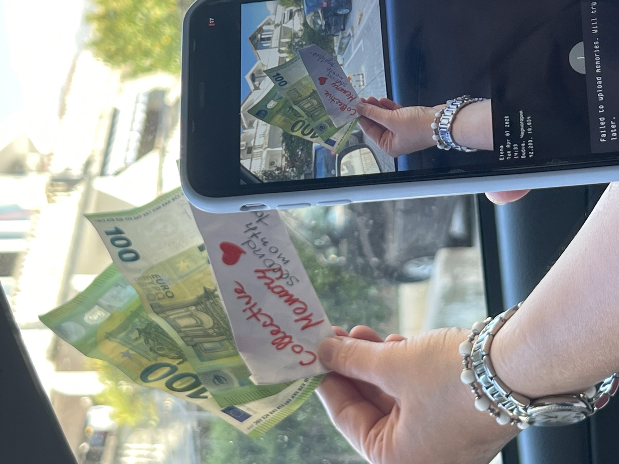A person is holding two 100 Euro banknotes and a handwritten note reading "Collective Memory Second month together" with a red heart drawn on it. They are inside a vehicle and taking a photo of the scene with their phone. The phone screen shows the same scene with the banknotes and note. The phone display also shows the date and time, "Tue Apr 07 2026 14:33," and the location, "Budva, Черногория 42.209, 10.034." The phone also displays the message "Failed to upload memories. Will try later." The person is wearing a silver bracelet-style watch on their left wrist. Outside the car window, one can see blurry buildings and cars.