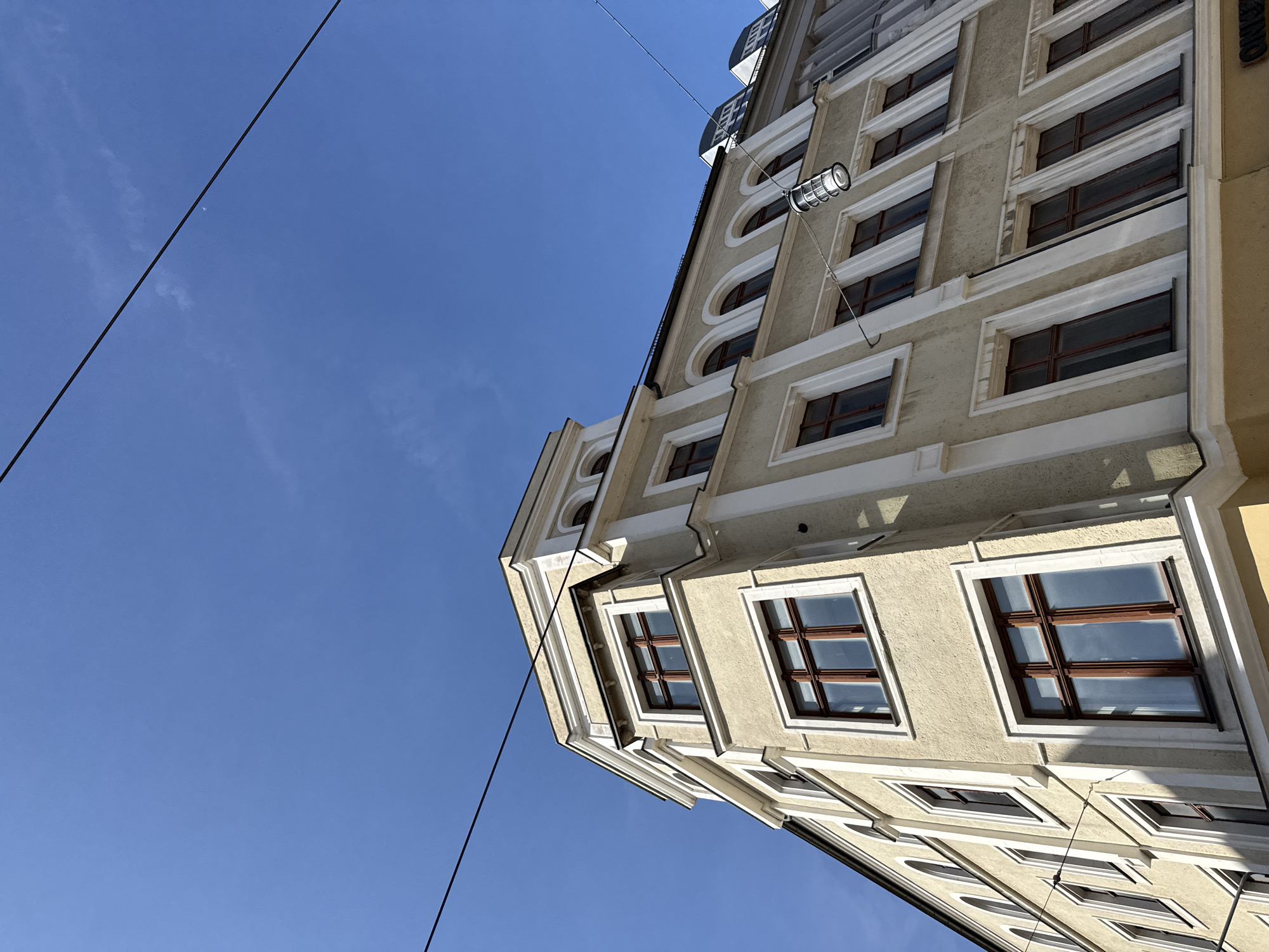 The image captures a low-angle view of a pale yellow, multi-story building in Görlitz, Germany, under a clear blue sky. The building features a distinctive corner design with several windows, each framed by white trim. The windows themselves have dark brown wooden frames and contain reflections of the sky. Some windows are rectangular, while others on the upper floors have arched tops.
The architecture suggests a historic European city. A thin black wire stretches across the top portion of the image, and another is visible diagonally across the building. A modern-style street lamp hangs from the diagonal wire in front of the building. The building's facade appears to be stucco, with decorative white molding around the windows and along the corners. The sunlight casts shadows on the building, indicating it is daytime. The scene is devoid of people, vehicles, or any immediate activity.
