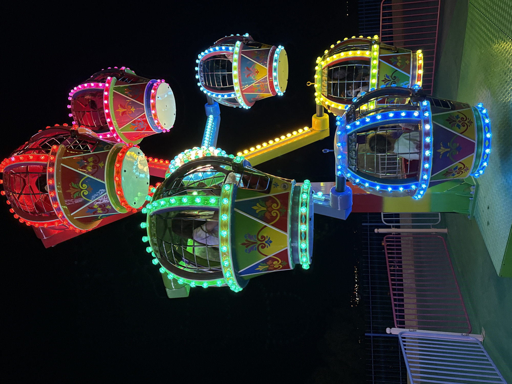 A brightly lit Ferris wheel at night, likely in a fairground or amusement park in Bodrum, Turkey, is the subject of this image. The Ferris wheel has multiple gondolas, each designed to resemble hot air balloons. These gondolas are adorned with colorful patterns, including geometric shapes and floral motifs, and are illuminated by strings of LED lights in various colors: red, pink, green, blue, and yellow. Several gondolas contain passengers, including what appears to be children. The Ferris wheel structure itself is a mix of yellow and blue metal. In the background, faint lights suggest a city or town, and a dark, silhouetted landscape. The foreground shows a green platform and fencing.