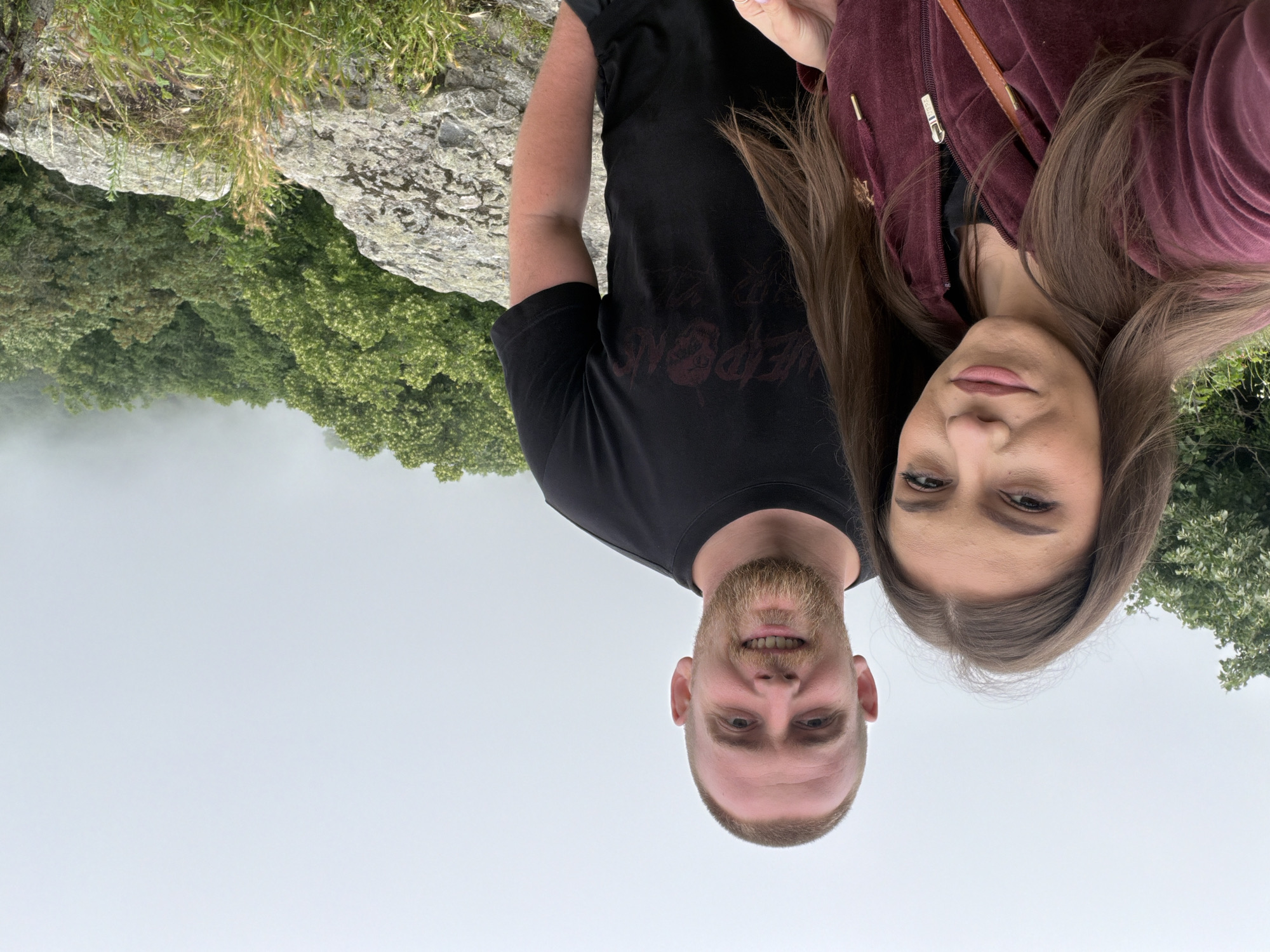 A portrait-style image features two individuals, a man and a woman, posing outdoors. The man is positioned on the left side of the frame, and the woman on the right. Both are looking directly at the camera.
The man has short brown hair and a short beard. He is wearing a black t-shirt with a faint, dark red graphic on the chest that appears to be text, possibly "WEAPONS," though it is not distinctly legible. The woman has long brown hair and is wearing a maroon zippered jacket. Small golden embellishments, possibly a logo resembling a polo player, are visible near the zipper of her jacket, but they are not clearly identifiable.
They appear to be standing on a rocky outcrop or cliff edge. To their left and below them, a dense forest of green deciduous trees stretches out. Further in the background, a thick layer of mist or fog partially obscures the view, creating a hazy, light gray and white horizon. The sky above is overcast, contributing to the diffused light in the scene.
Both individuals display neutral to slightly smiling expressions. The man has a broader smile, showing his teeth, while the woman's smile is more subtle and closed-mouthed. The overall impression is that of a couple taking a photograph together at a scenic overlook, possibly in a mountainous or hilly region, on an overcast or misty day.